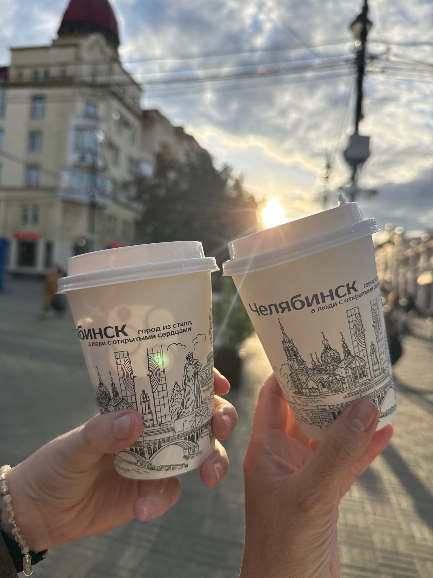 Two paper coffee cups, each adorned with a black and white sketch of urban architecture and text that reads "Челябинск город из стали, а люди с открытыми сердцами" (Chelyabinsk, a city of steel, and people with open hearts), are held up by two hands in the foreground. The cups are tilted towards each other, appearing to be clinked in a toast. The background is a blurred cityscape, featuring buildings and a street lamp with overhead wires. The sun is setting, casting a warm, golden glow across the scene and creating lens flare. The overall impression is of a casual, urban moment shared outdoors.