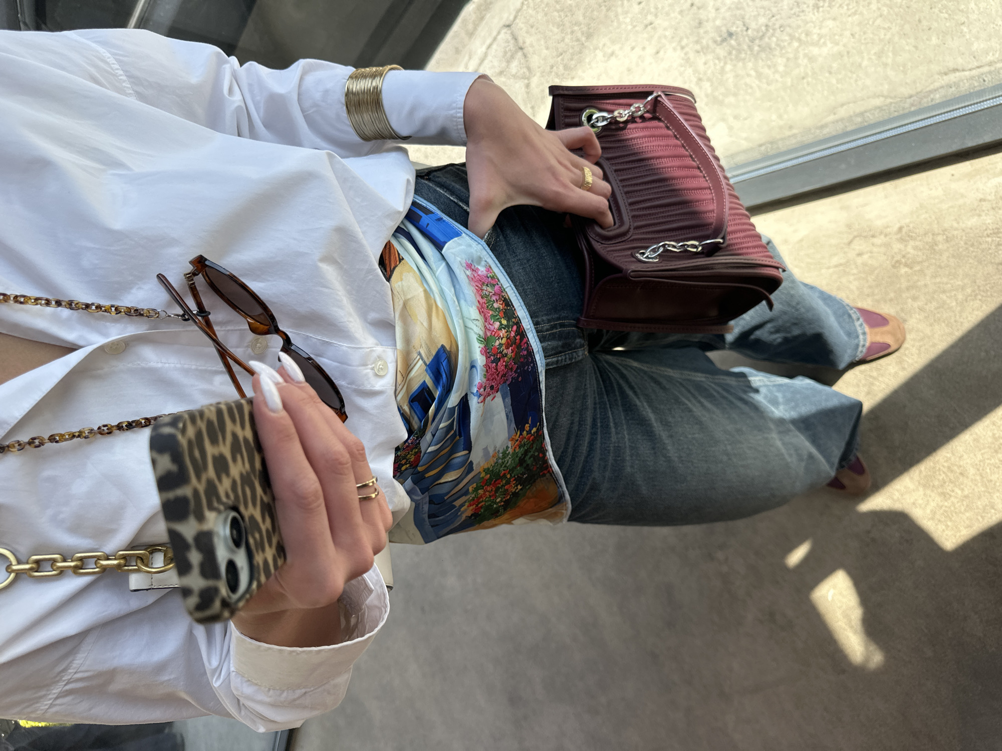 The image provides a high-angle, close-up view of a person seated outdoors in Yerevan, Armenia. The subject is wearing a white long-sleeved shirt and blue jeans. A colorful silk scarf, featuring a detailed print of a scenic European-style town with buildings and vibrant flowers, is tucked into the waistband of the jeans.
In the person's left hand, a smartphone with a leopard print case and a thick gold chain strap is held. The right hand rests on a maroon or burgundy handbag detailed with silver chains. The person's wrists are adorned with stacked gold bangles on the right, and gold rings are visible on both hands, which have white-painted nails. A pair of brown sunglasses, attached to a tortoiseshell-patterned chain, are draped over the white shirt. One foot, wearing a brown shoe with a purple top, is partially visible.
The scene is brightly illuminated by direct sunlight, casting distinct, sharp shadows on the light-colored, textured ground, indicative of a clear, sunny day, likely mid-morning or afternoon. A dark, linear element in the background suggests a structural feature like a window frame or railing. The activity depicted is casual, with the person appearing to be sitting and holding their belongings. No visible text is present.