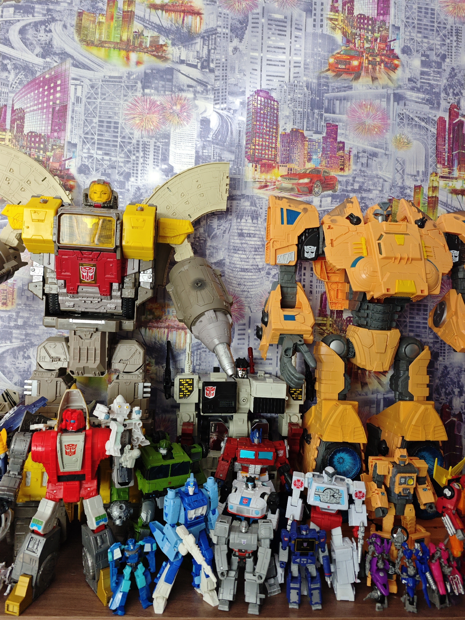 The image shows a collection of Transformers toys, mostly Autobots, arranged on a wooden shelf against a backdrop of a cityscape wallpaper with overlaid imagery including cars, bridges, and fireworks. Prominent figures include a large yellow and gray Transformer with a red chest symbol on the left, an orange Transformer on the right, and a white and black Transformer standing in the center. Other Transformers of varying sizes and colors are clustered in front, including red, green, blue, and purple figures. The photograph is taken indoors with the focus on the toys. There's no evidence to suggest the location is Markovskii, Russia.