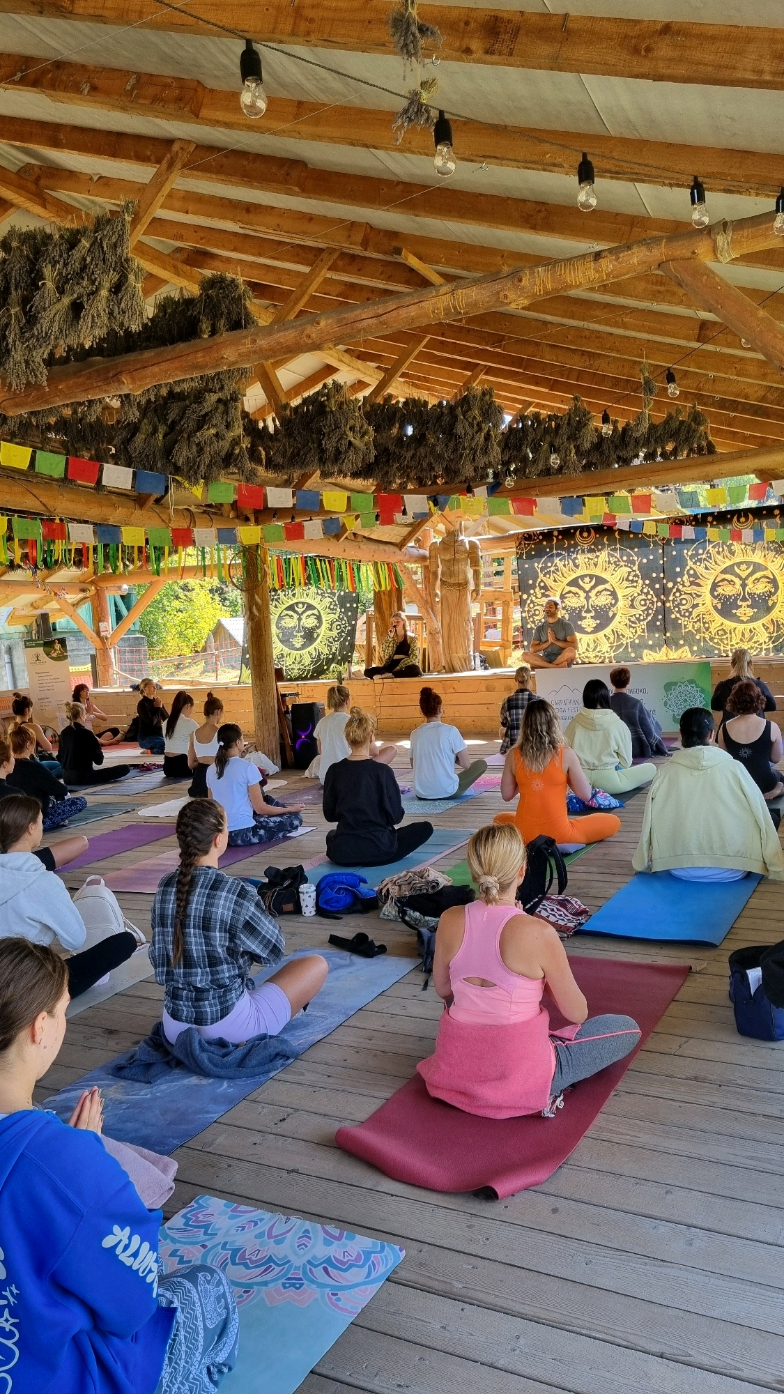 The image captures an outdoor yoga session under a wooden, open-air structure. Numerous people, primarily women, are seated on yoga mats arranged on a wooden floor, facing a raised platform. On the platform, two individuals appear to be leading the session. One person, dressed in dark clothing, sits cross-legged, while another, in a lighter colored shirt and pants, stands and gestures. Behind them, a backdrop features a large, ornate sun symbol against a dark fabric. Prayer flags in various colors are strung across the beams of the structure, suggesting a spiritual or cultural event. Bunches of dried herbs, likely lavender, hang from the ceiling, adding to the rustic ambiance. The scene is well-lit, indicating it is daytime, and the natural light filtering through suggests fair weather. The overall atmosphere seems peaceful and focused, with participants engaged in meditation or yoga practice. In the background, trees and greenery are visible, placing the setting in a natural environment. A sign with Ukrainian text is partially visible on the left side of the image.