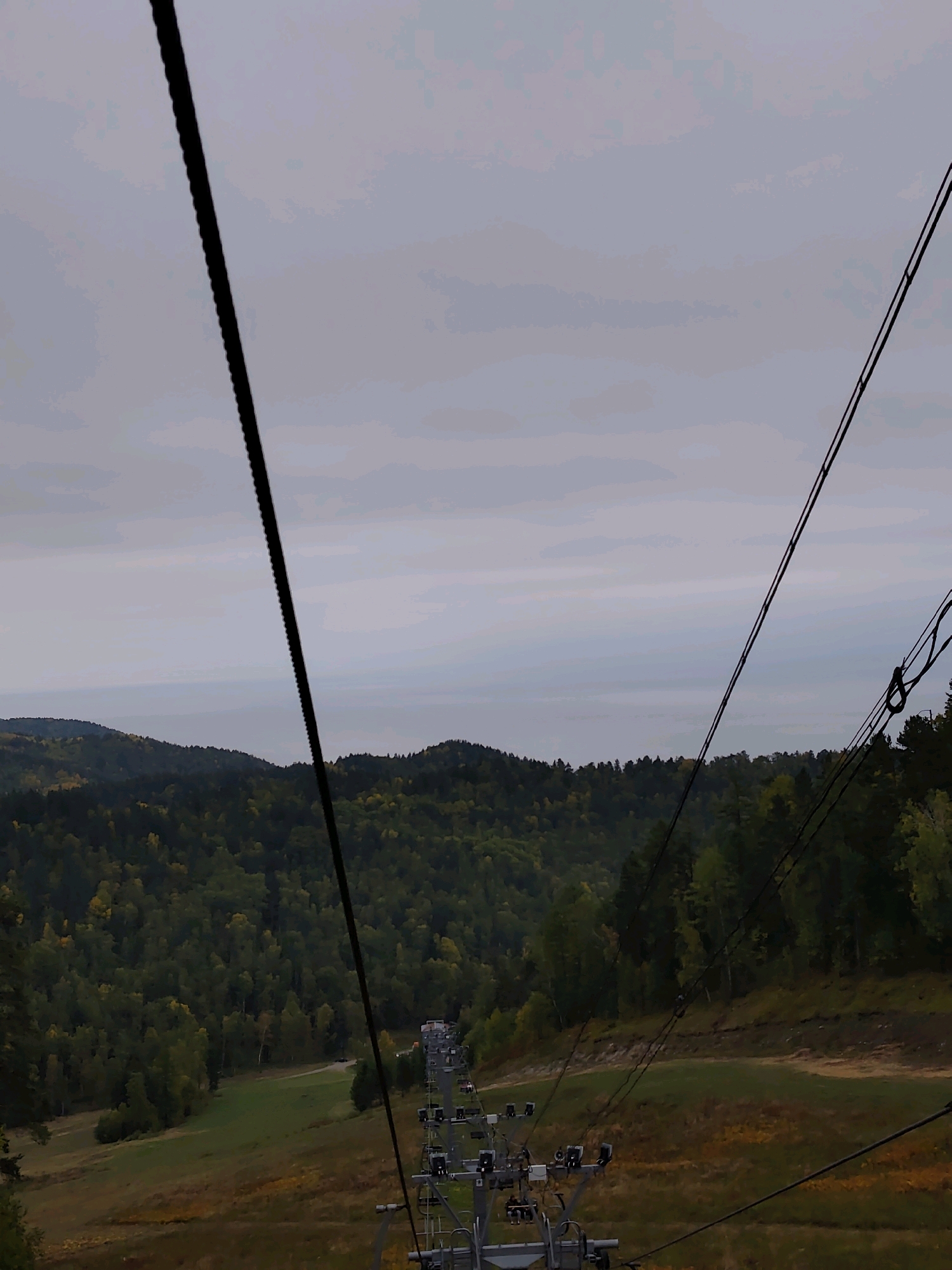 This is a view from a ski lift ascending a mountain slope. The ski lift cables are prominently in the foreground, stretching diagonally across the frame. Below, a lush green forest covers rolling hills, with scattered patches of yellow indicating autumn foliage. A grassy slope with some autumnal colors is visible on the right side of the image, and a ski lift structure with chairs is also present on the right. In the distance, a hazy body of water, possibly a lake or sea, is visible beyond the hills, under a cloudy sky. The location is the city of Nikola, Russia, specifically on a ski resort within the city. The scene captures a scenic natural environment during the transition to autumn.