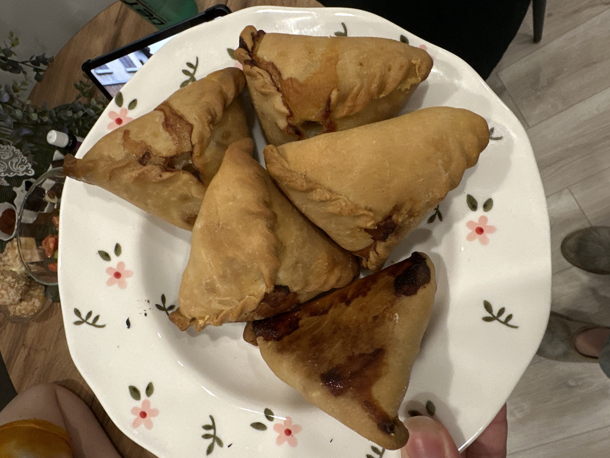 The image shows a plate of five fried pastries resembling samosas. The plate is white with a floral design of pink flowers and green leaves. One pastry is significantly darker than the others, appearing overcooked. The plate is being held in someone's hand. Background elements include a wooden table, a tablet device, a glass container with assorted foods, and a glimpse of flooring that could be hardwood or laminate. There are no clear indicators in the image to suggest the city of Kazan, Russia.