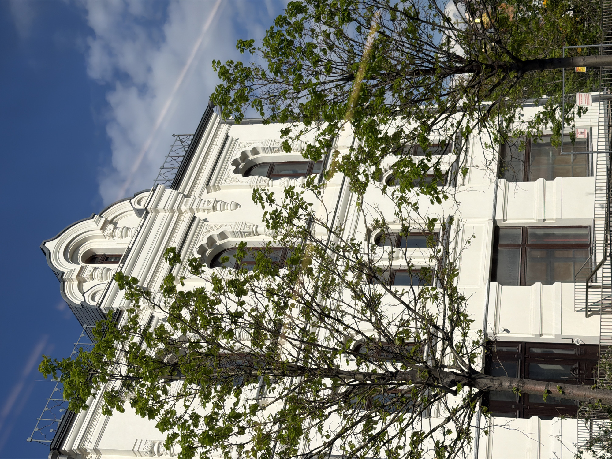 A tilted, medium shot reveals a white building in Moscow, Russia, partially obscured by two trees with green leaves. The building has a distinct architectural style, featuring ornamental designs, arched windows with brown frames, and white facade elements. A metal fire escape is visible on the right side of the building. The sky, a clear blue with scattered white clouds, forms the background, accented by a faint white streak.