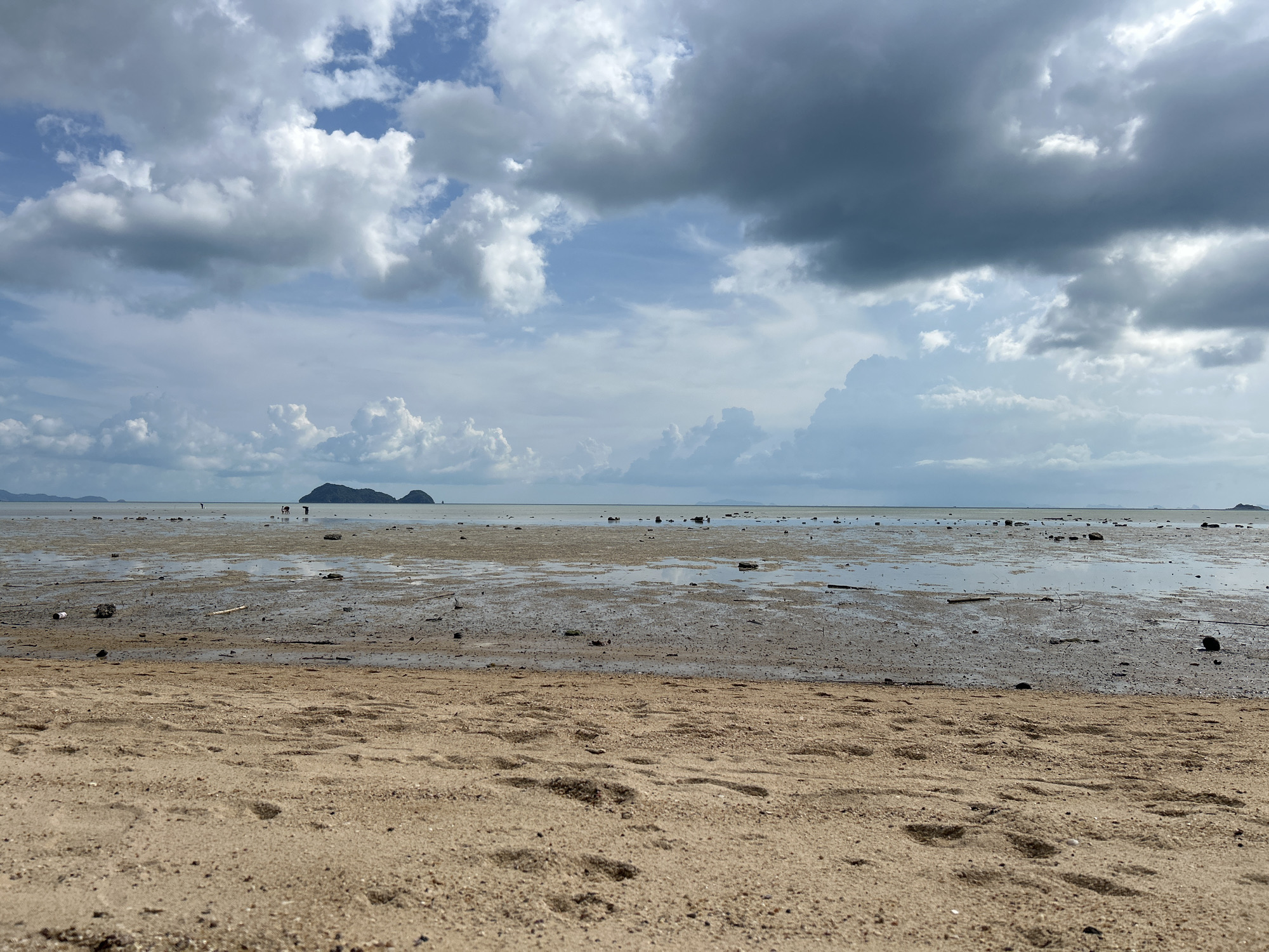 The scene is a beach at low tide in Baan Sri Thanu, Thailand. The foreground is composed of light-brown sand with visible footprints. The middle ground shows a wide expanse of muddy tidal flats with scattered debris and shallow pools of water. In the distance, a pair of small, green islands are silhouetted against the horizon. Several human figures are visible on the tidal flats, appearing small due to the distance. The sky is dominated by a mix of white cumulus and dark, grey storm clouds.