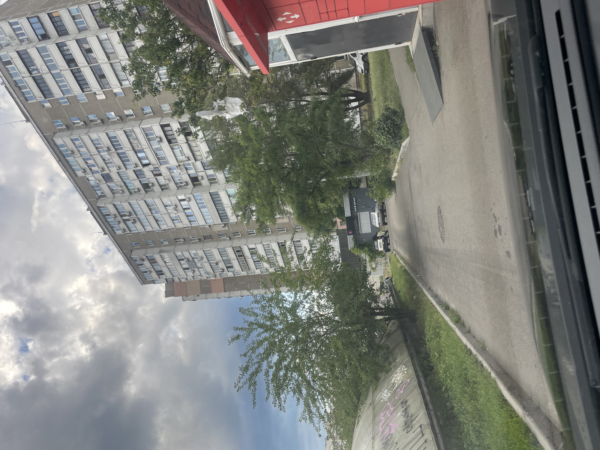 The image captures a street scene in Kyiv, Ukraine, under a cloudy sky. A large, multi-story residential building dominates the background, with numerous windows and air conditioning units visible. In the foreground, a paved road leads away from the viewer, flanked by grassy areas and trees. A large, curved, graffiti-covered structure is on the left side of the road. On the right, a red structure with a dark door and a ramp extends towards the road. Several cars are parked or driving on the road. The scene suggests a typical urban environment in Kyiv.