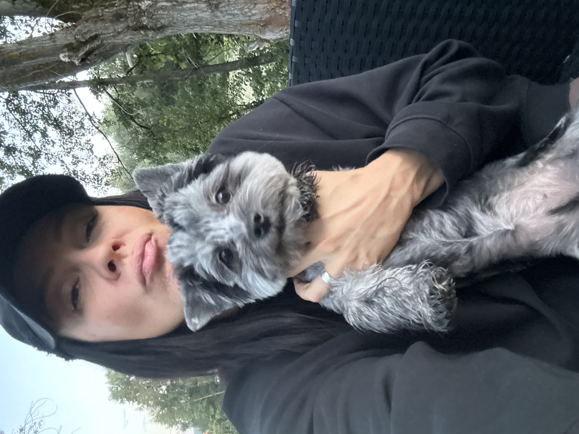 A medium shot, outdoors, shows a woman with dark hair and wearing a black baseball cap and black sweatshirt holding a small, gray and black dog. The woman is puckering her lips as if to kiss the dog. The dog is looking towards the camera. The background consists of green trees and a light-colored sky, suggesting it is daytime. The woman has a silver ring on her left ring finger. The scene appears to be a park or backyard in Chaikovsky, Russia.