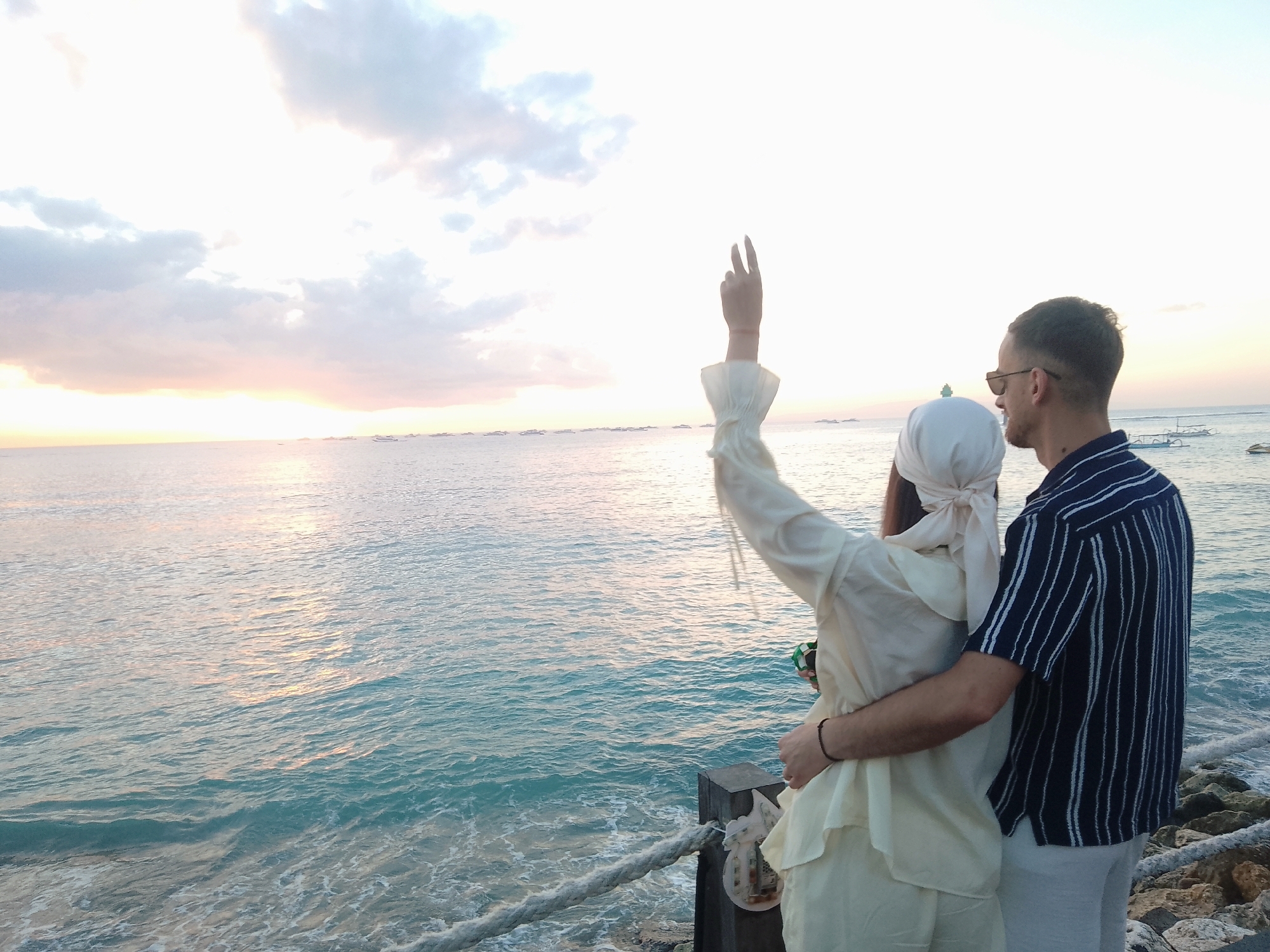 A couple stands on a rocky shore overlooking the ocean at sunset in Denpasar, Indonesia. The woman, on the left, wears a white headscarf and a cream-colored outfit with ruffled sleeves, and she raises her right arm in a V-sign gesture. The man, on the right, has short hair, wears sunglasses, and a dark blue striped short-sleeved shirt. He has his arm around the woman's waist. The ocean is a turquoise blue with small waves and reflections of the sunset sky. The sky is mostly white with some soft pink and orange hues near the horizon, and there are several boats visible in the distance. A thick rope railing runs along the edge of the shore in the foreground.