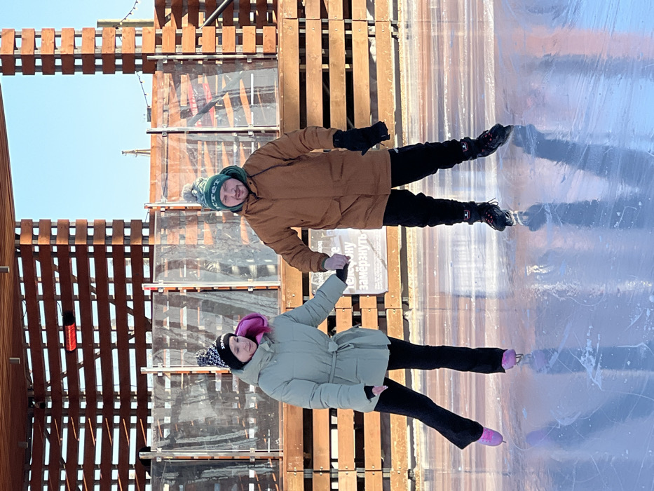 An outdoor scene captures two individuals ice skating on a bright, reflective ice rink. The location is an ice rink at New Holland Island in Saint Petersburg, Russia.
The subject on the left is a female, wearing a light sage-green belted puffer jacket, black flared pants, and black ice skates with pink blades and laces. She has a black knit beanie with white Cyrillic lettering and a prominent pink tuft of material or hair visible on her left side.
The subject on the right is a male, wearing a brown winter parka, black pants, black gloves, and black ice skates. He wears a green knit beanie with white Cyrillic lettering.
Both individuals are actively ice skating and holding hands, with the male's gloved left hand clasping the female's gloved right hand. Their posture suggests forward motion.
In the background, a multi-tiered wooden structure with horizontal slats borders the rink. The lower section features a transparent barrier above a solid wooden base. A red rectangular infrared heater is mounted on the upper wooden structure to the left. A white sign with black Cyrillic text, partially legible as "...телефонy замерзнуть" (approximately "...to freeze the telephone"), is visible behind the male subject. The sky above is clear and bright blue, indicating sunny weather.
