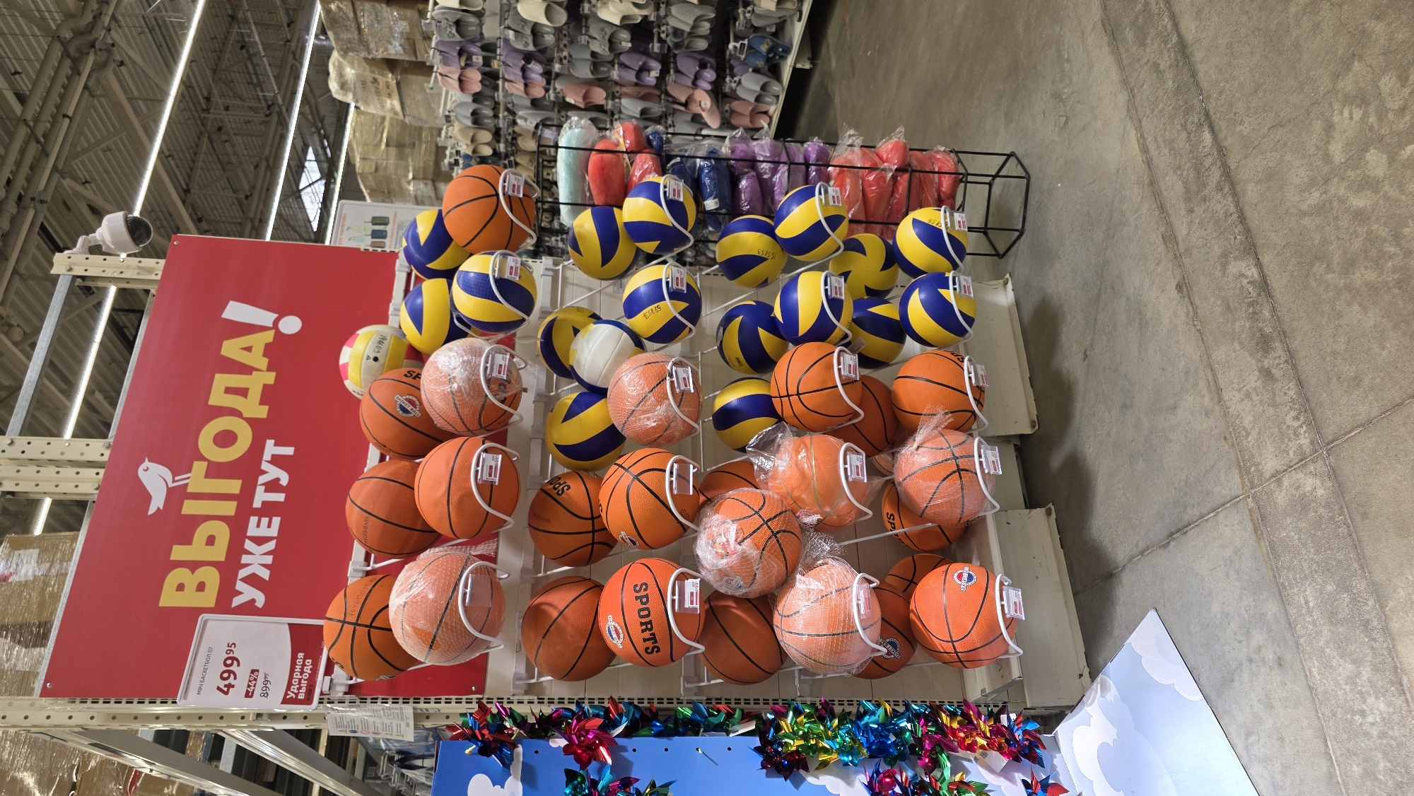 This image captures an indoor retail store environment, primarily featuring a display of sports balls. The main subjects are numerous basketballs and volleyballs, neatly arranged on white metal racks. The basketballs are mostly orange, some still enclosed in plastic netting, with brand names like "SPORTS" or "SP" visible. The volleyballs are predominantly blue and yellow, with some featuring the numbers "155859".
To the left, a large, vibrant red sign reads "ВЫГОДА! УЖЕ ТУТ" (BENEFIT! ALREADY HERE) in yellow Cyrillic script, accompanied by a small bird emblem. Attached to this sign is a white price tag detailing "МЯЧ БАСКЕТБОЛ 07" (BASKETBALL BALL 07) for "499.95", indicating a "44%" discount from an original price of "899.99", and labeled as "Ударная выгода" (Shocking benefit/deal).
In the background, shelves are stocked with various other items, including rows of colored slippers or sandals. The setting features a high ceiling with visible structural elements and overhead lighting, suggesting a large commercial space. A white security camera is mounted high on a support beam in the upper left. The floor is a light grey, textured concrete. The bottom of the display is decorated with colorful, metallic pinwheels or frills. The scene depicts a merchandising setup within what appears to be a general goods or department store. No people are visible in the image.