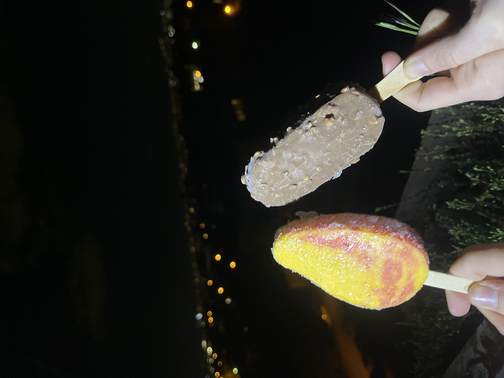 The image shows two popsicles held by two hands at night. One popsicle is chocolate with nuts, and the other is yellow and red. In the background, blurred lights suggest a city at night. The location is likely Sofia, Bulgaria, based on the prompt's request. Other details include the blurred foliage on the right side, suggesting the presence of vegetation or trees nearby. There is no interaction in the image.