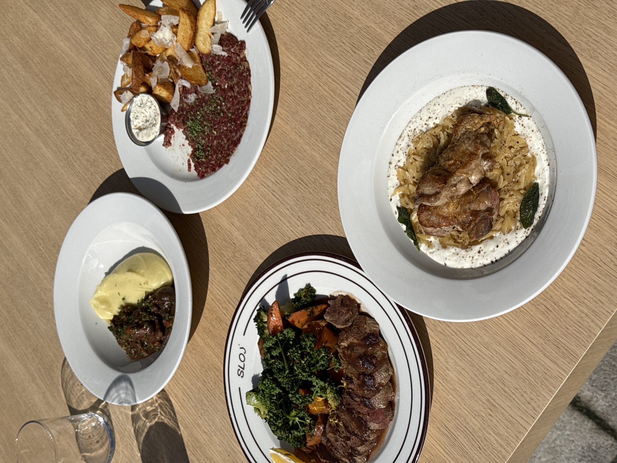 This top-down image showcases four distinct gourmet dishes laid out on a light wooden table, likely during a daytime meal. Strong, dark shadows cast by the plates and a glass indicate bright sunlight, suggesting an outdoor setting or a well-lit indoor space.
The top left plate features a generous serving of finely minced steak tartare, partially garnished with green herbs. It is accompanied by golden-brown potato wedges or thick-cut fries, sprinkled with grated cheese (possibly Parmesan), and a small metal cup of creamy dipping sauce.
On the top right, a dish presents two pieces of seared or roasted meat, possibly chicken or pork, resting on a bed of orzo pasta or rice mixed with sautéed onions. A creamy white sauce covers the bottom of the plate, and fresh sage leaves adorn the dish.
The bottom left plate holds a smaller serving of smooth, yellow mashed potatoes or a similar puree alongside a darker, rich-looking stewed meat dish, garnished with green herbs.
The bottom right plate is a distinctive oval design with brown and white striped borders and the word "SLOJ" printed in black on the left side. It contains slices of medium-rare seared meat, likely steak, drizzled with a sauce. This is served with vibrant green broccoli florets, roasted orange root vegetables (possibly carrots), and a lemon wedge.
A clear glass tumbler is partially visible in the bottom left corner. The scene depicts a diverse culinary spread, suggesting a pleasant dining experience in Belgrade, Serbia.