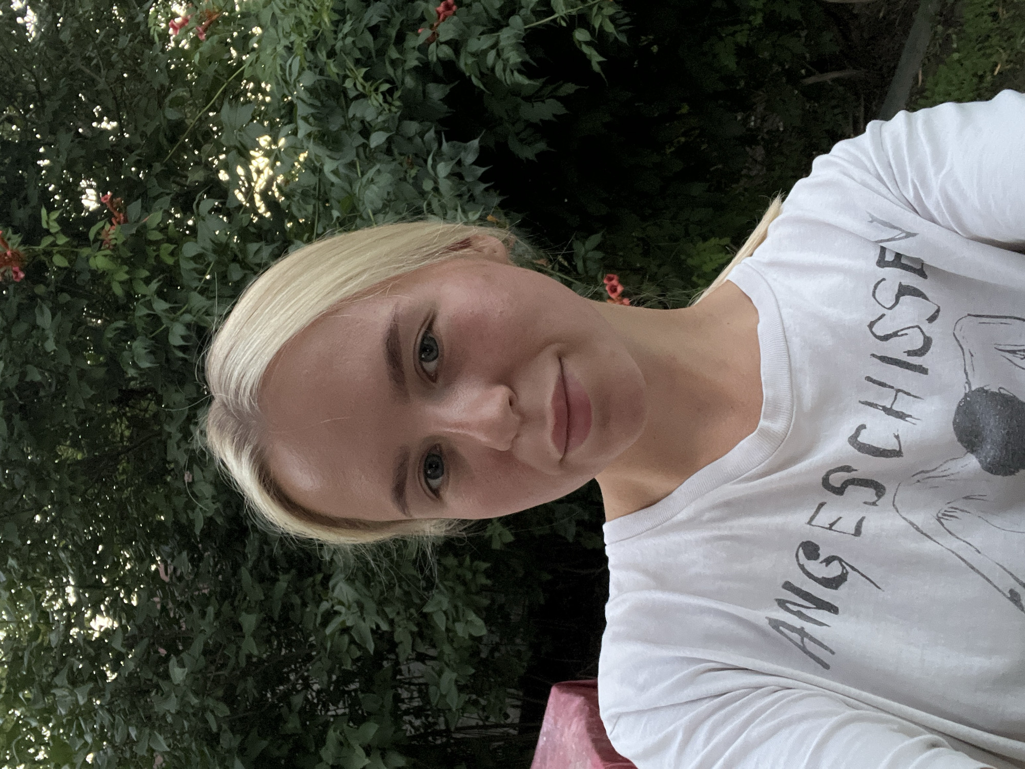 An outdoor, eye-level, medium shot shows a young blonde woman with fair skin and blue eyes taking a selfie. She is looking directly at the camera with a neutral expression. She has on a white t-shirt with a graphic print and some dark text that appears to read "ANGESCHISSEN". Behind her, there is a dense green bush with some red flowers visible. The background is slightly blurred, suggesting the photo was taken with a shallow depth of field. The lighting is natural, and it appears to be daytime, possibly late afternoon or early evening, as there is a soft glow filtering through the leaves of the bush. To the left of the woman, a portion of a red-covered table is visible. The overall scene suggests a casual, outdoor setting, possibly a backyard or garden.
