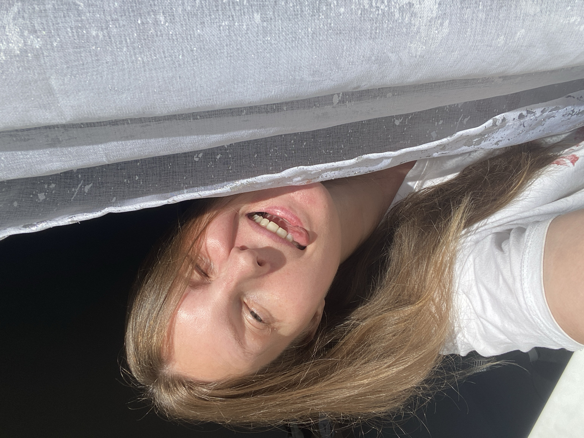 A woman with long brown hair is smiling with her tongue slightly protruding. She is wearing a white shirt. Above her is a white, semi-transparent, textured fabric. The background is dark. The location in the city of Noviy, Russia, cannot be determined from the image.
