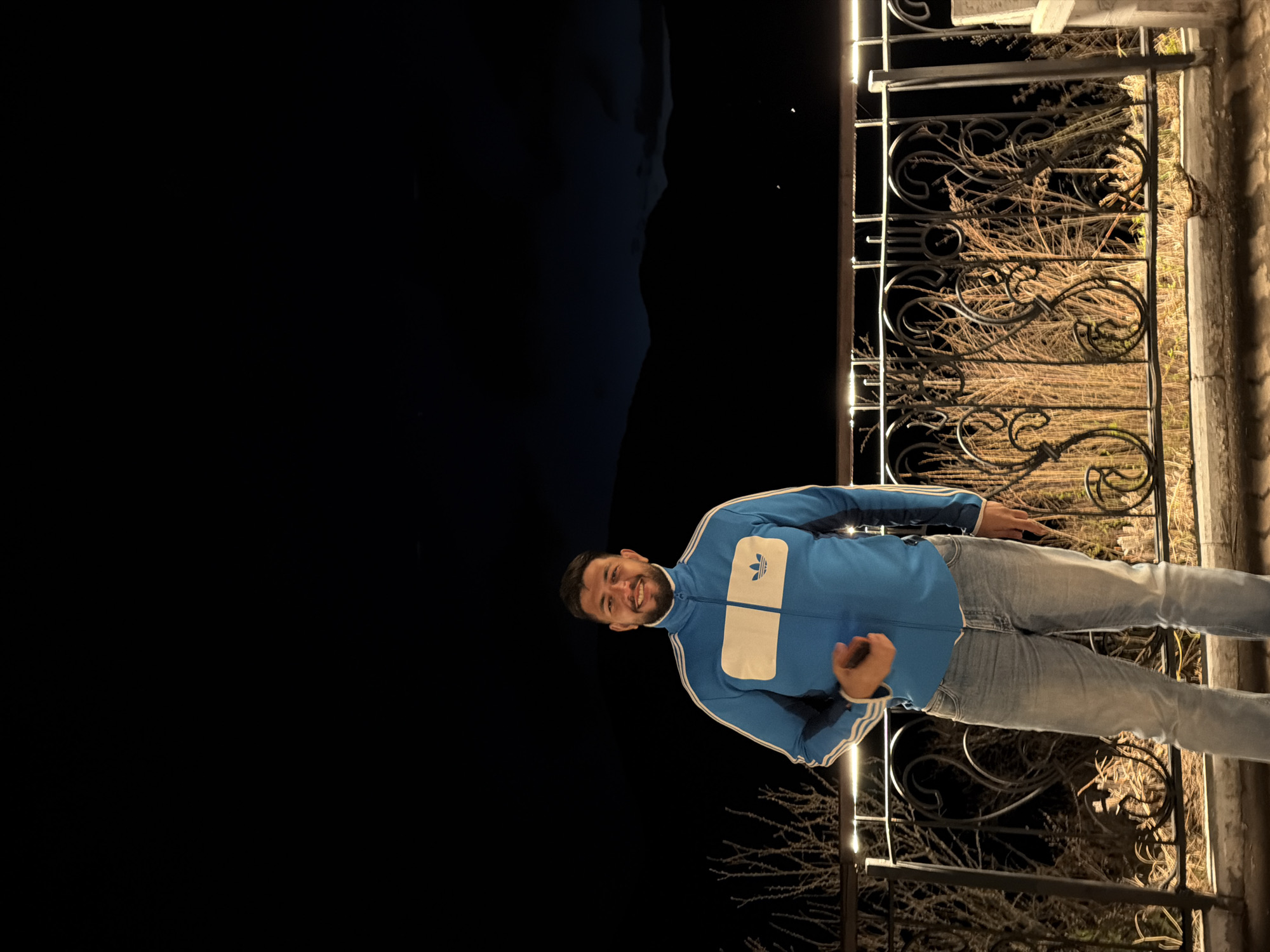 A man stands on a porch with a wrought iron railing in Sliznevo, Russia. He is wearing a blue Adidas track jacket and jeans. Behind him is a dark landscape.