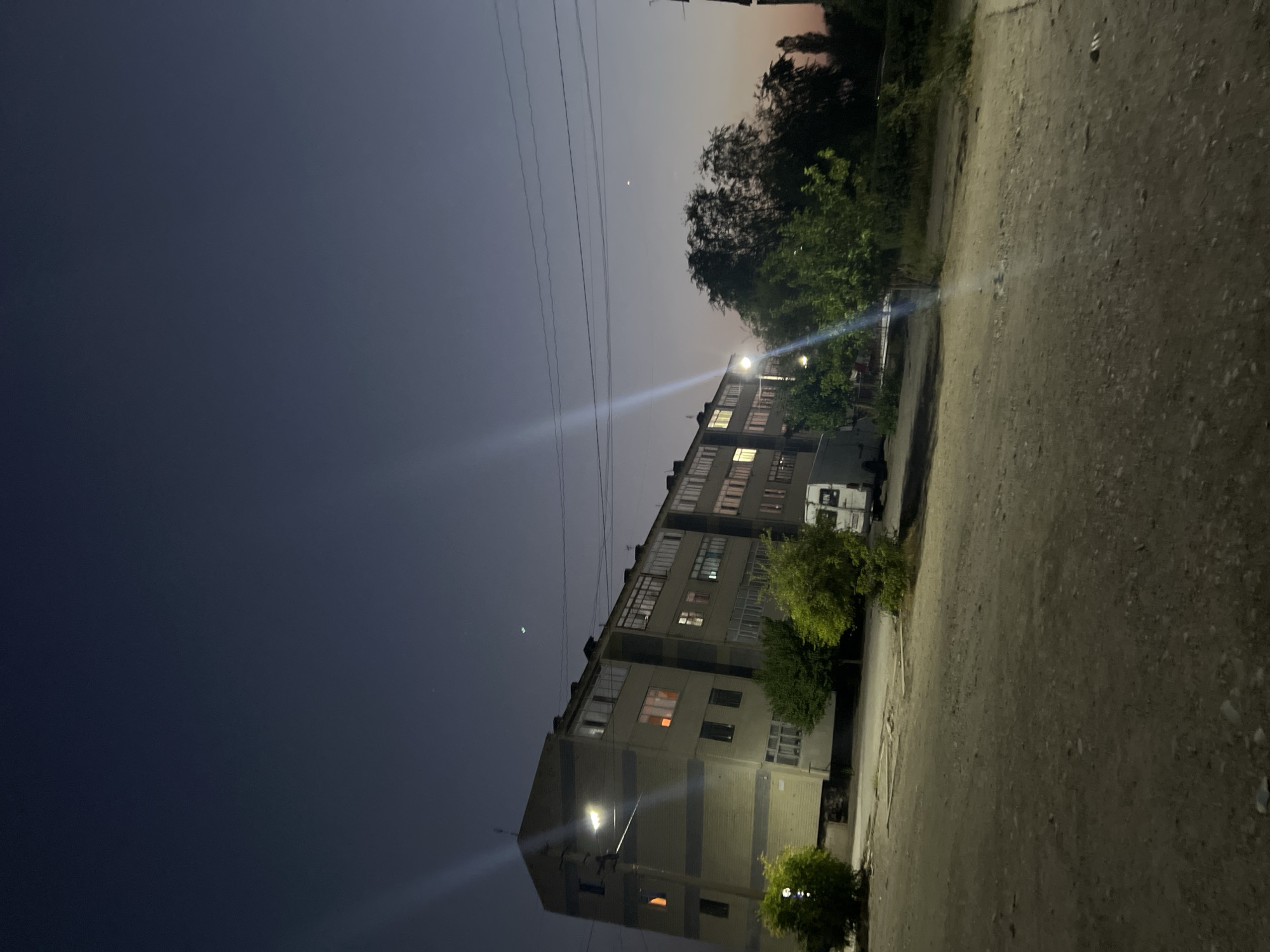 The image depicts a street scene in Şamaldı-Say, Kyrgyzstan, during twilight. A multi-story apartment building dominates the left side of the frame, its windows illuminated with warm light, suggesting occupancy. A streetlamp casts a bright glow on the building's facade. Green trees are scattered along the sidewalk in front of the building, and a white van is parked to the right. The foreground is a dirt road or unpaved area with visible gravel and debris. The sky is a deep, dusky blue, with thin power lines visible against it. There are no people visible in the image.