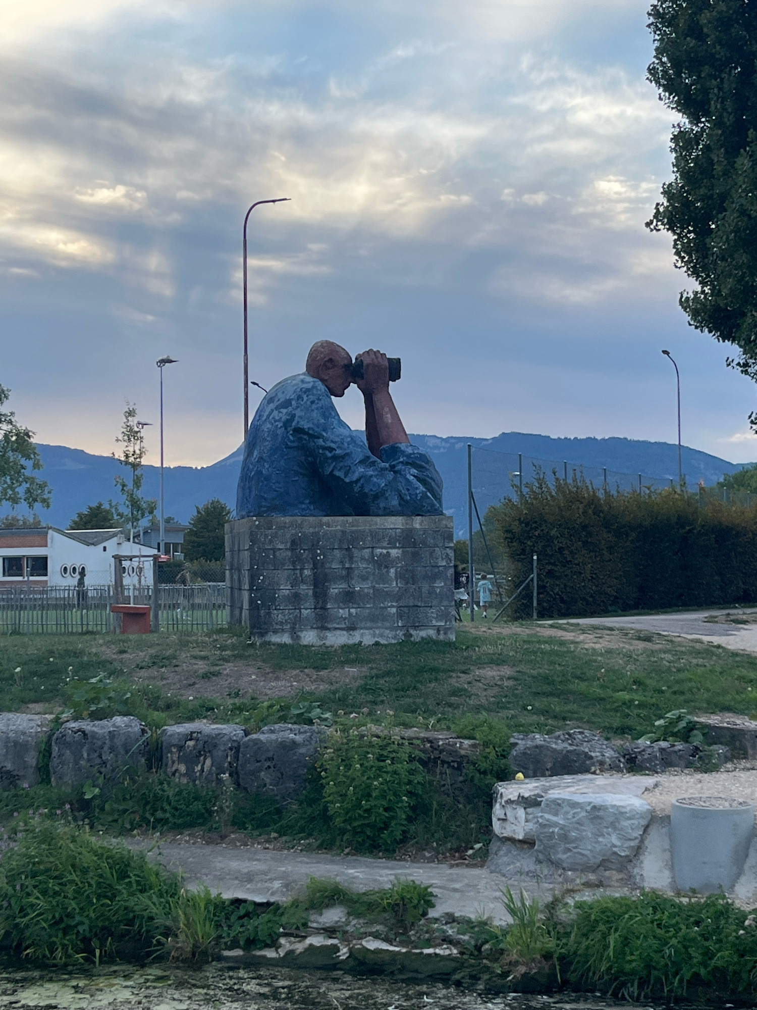 This is a photograph of a large, seated sculpture of a person looking through binoculars. The sculpture is colored blue and is positioned on a grey, block-like base. The sculpture is situated outdoors in what appears to be a park or recreational area.
In the background, there is a building with a white facade and red trim, featuring several circular windows. A fence encloses an area behind this building, and beyond that, a line of dense green bushes is visible. In the far distance, a range of blue-hued mountains rises against a cloudy sky. Several lampposts are present in the scene.
The foreground features grass, stones, and a body of murky water. The overall atmosphere suggests an overcast day.
This sculpture is located at the Parc des Rives in Yverdon-les-Bains, Switzerland.