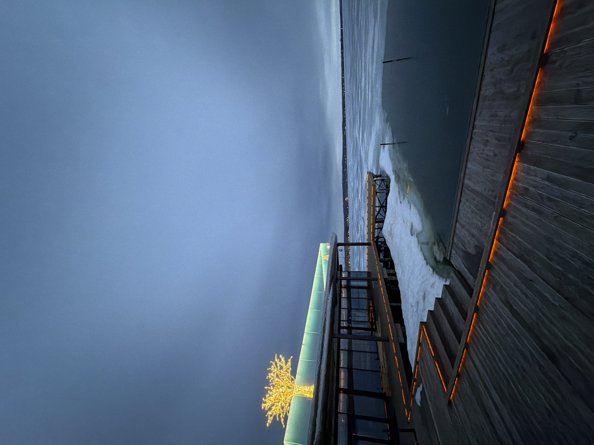 The image depicts a winter scene near the embankment of the Votkinsk Reservoir in Votkinsk, Russia. A building with a glass facade adorned with festive yellow lights is visible on the left. To the right, there's a wooden boardwalk or deck with integrated orange lighting. The reservoir is partially frozen, with ice and snow covering the surface near the shore. In the distance, the lights of the city line the horizon. The sky is a dark, overcast blue.