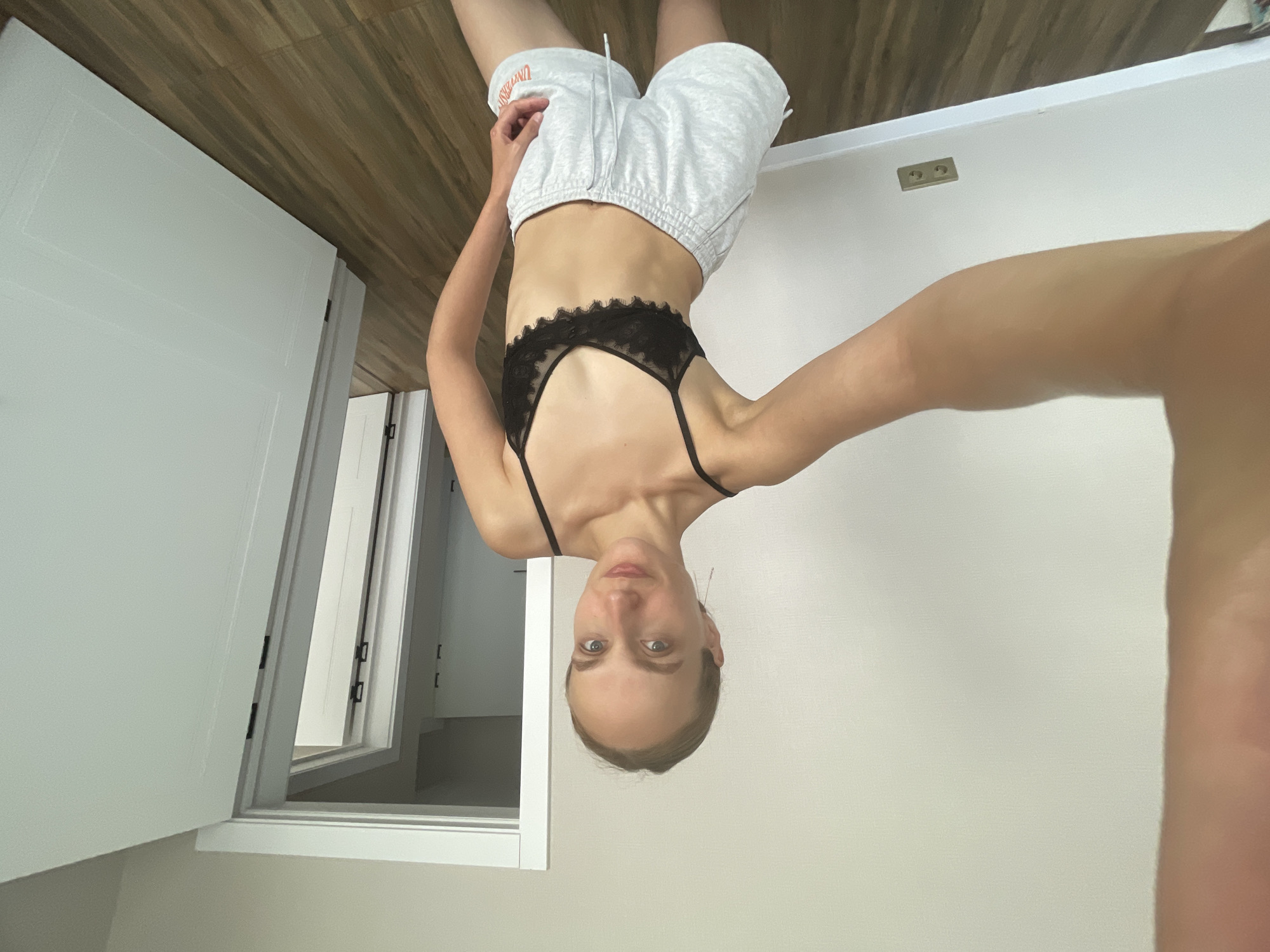 A woman with blonde hair pulled back stands in a room, taking a selfie. She is wearing a black lace bra and light grey athletic shorts. The room has light-colored walls and dark wood flooring. In the background, there are several interior doors, one of which is partially open, revealing another doorway beyond. The overall impression is that of a casual, indoor setting. No specific location in Podshivalovo, Russia, can be identified from the visual information provided.