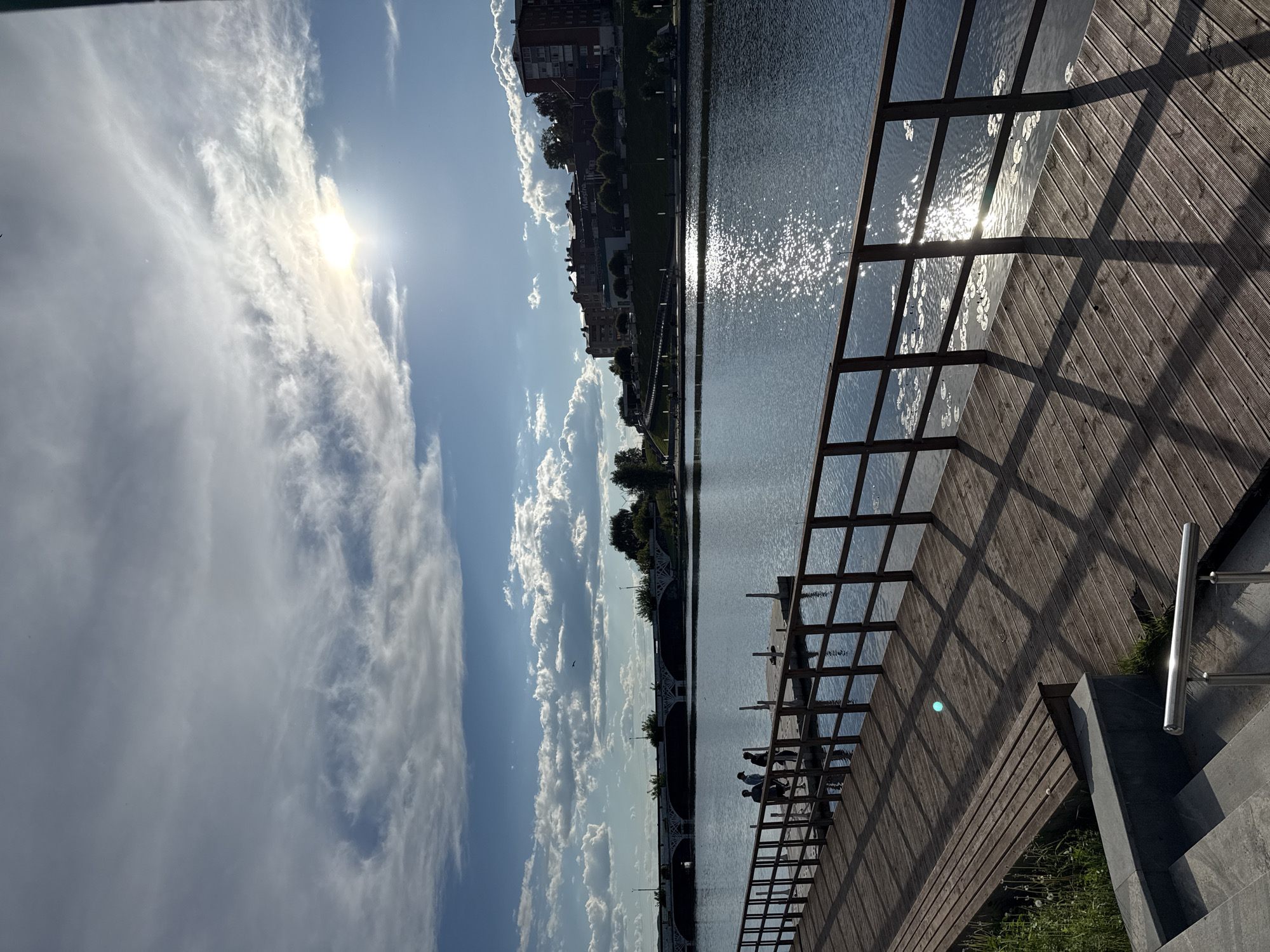 When viewed in landscape orientation, the image displays a bright, sunny daytime scene along a waterfront in Votkinsk, Russia. In the foreground, a wooden boardwalk extends along the edge of a wide body of water. A wooden railing creates prominent, grid-like shadows across the boardwalk due to the intense sunlight. Towards the right, a modern concrete staircase with a metal handrail is visible. Several silhouetted figures are seated or lounging on the wooden structure next to the water, appearing to relax and enjoy the setting.
The midground features the expansive water, which reflects the brilliant sun, creating numerous sparkling glints across its surface. A multi-arched bridge spans the water in the distance. The far bank is lined with a town or city, characterized by multi-story buildings and areas of greenery. No discernible text is visible on these distant buildings.
The background consists of a bright blue sky adorned with scattered, wispy white clouds. The sun, a powerful light source, is visible high in the sky, partially veiled by clouds, causing a noticeable lens flare and illuminating the entire scene. The overall impression is one of a pleasant, leisurely day with clear weather and strong sunlight.