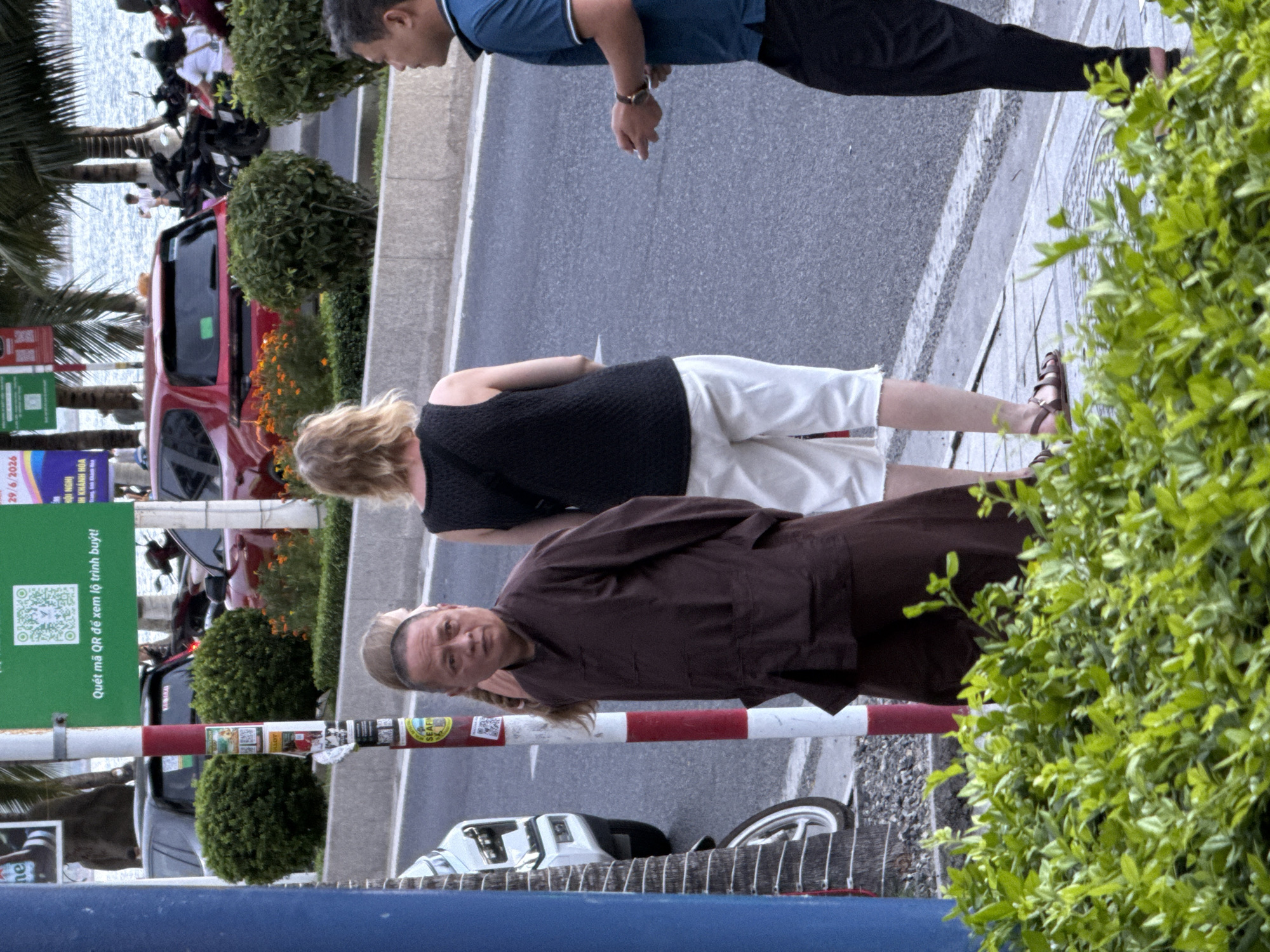 The image depicts a street scene in Nha Trang Ward, Vietnam, with a focus on three individuals on a sidewalk adjacent to a road. In the foreground, lush green foliage partially obscures the lower portion of the scene.
On the left, a person wearing a traditional brown monk's robe stands near a red and white striped pole. Their gaze is directed towards the right. Behind them, another individual with blonde hair is visible, wearing a black sleeveless top and white shorts. They are also facing towards the right, with their back to the camera.
To the right, a man in a blue polo shirt and black pants stands with his hands in front of him, holding something small. He is facing to his left, looking at the other individuals. His left arm is bent, and his fingers are partially extended.
The background shows a road with parked cars, including a red hatchback. Palm trees are visible in the distance, suggesting a tropical environment. A body of water, likely the sea, is also visible beyond the trees and a building. A green sign with a QR code and Vietnamese text is mounted on the pole next to the monk. Traffic lights and street lamps are also present in the background. The overall impression is a busy urban street scene.