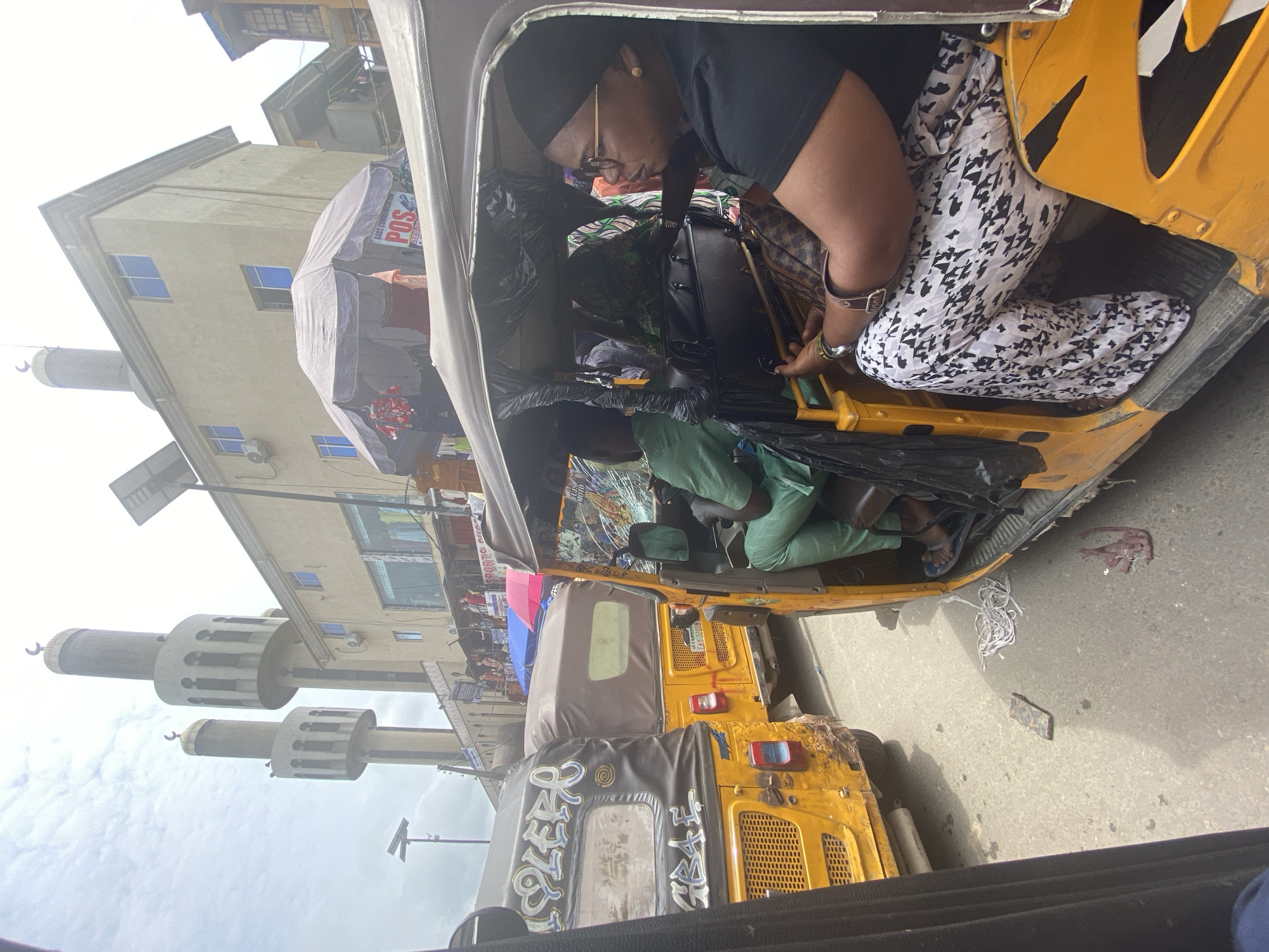 A street scene in Lagos, Nigeria, shows a traffic jam of yellow auto-rickshaws, known as "Keke NAPEPs." The vehicles are densely packed, with one auto-rickshaw in the foreground occupied by a woman wearing glasses, a black t-shirt, and patterned trousers, and a person in a green outfit driving. The auto-rickshaw has a cracked windshield. Another auto-rickshaw to the left displays graffiti. In the background, a building with two minarets, likely part of a mosque, stands against a cloudy sky. Shops and market stalls are visible along the street.
