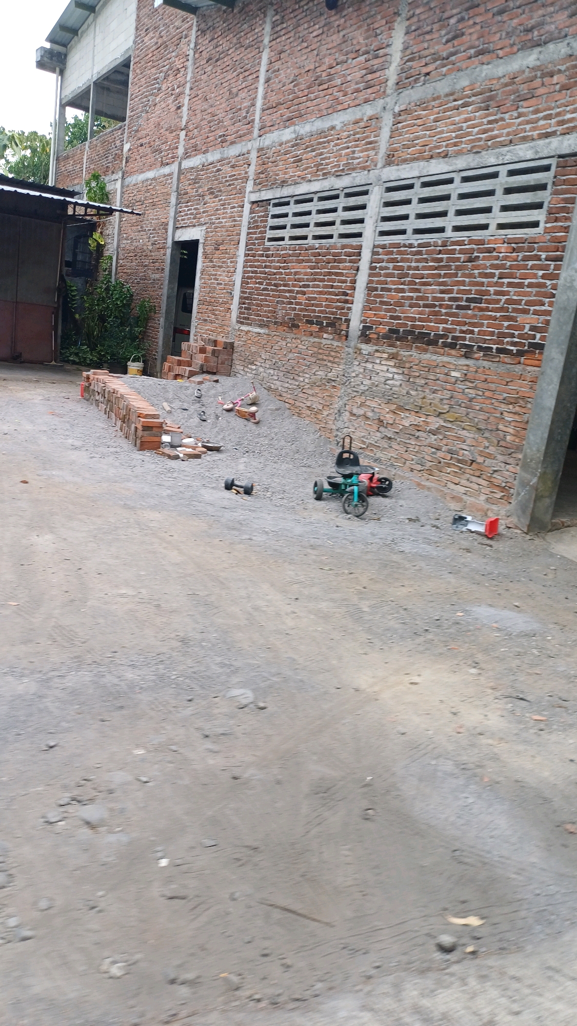 This is a ground-level, eye-level photograph taken outdoors at a construction site or storage area in Badran, Indonesia. The scene depicts a brick building with concrete support beams and ventilation openings. In the foreground, a dusty, gravelly ground is visible, with tire tracks suggesting vehicle movement. Several objects are scattered on the ground, including a pair of dumbbells, a small pedal-powered go-kart, and a red and white object that appears to be a traffic cone or warning sign. In the mid-ground, there is a pile of bricks and a heap of gravel, with some smaller scattered bricks and debris. Further back, near the building, a stack of bricks is neatly arranged, and a yellow bucket sits nearby. Green foliage and a covered structure are visible to the left of the building. The overall impression is one of an unfinished or in-progress construction environment.