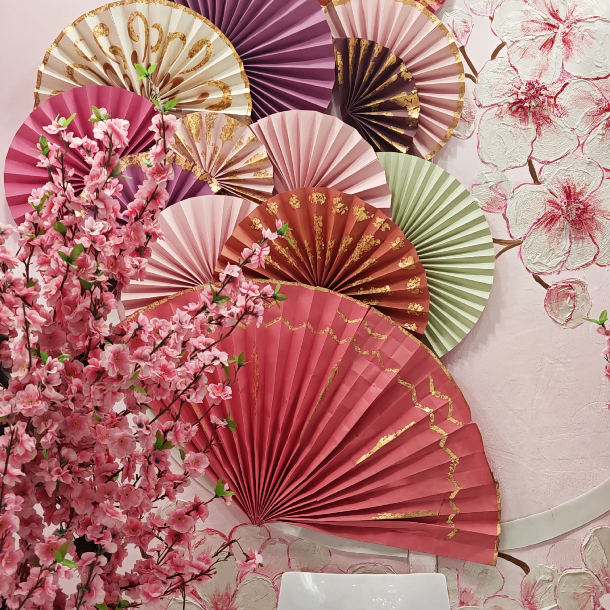 A close-up shot displays a decorative display with a pink theme. The background wall is adorned with embossed white and pink flower paintings. Arranged on the wall are numerous colorful paper fans, some decorated with gold accents. To the left, a cluster of pink cherry blossom branches adds depth to the composition. The setting might be a bathroom, as suggested by a part of a white sink visible at the bottom. The location is Taganrog City District, Russia.