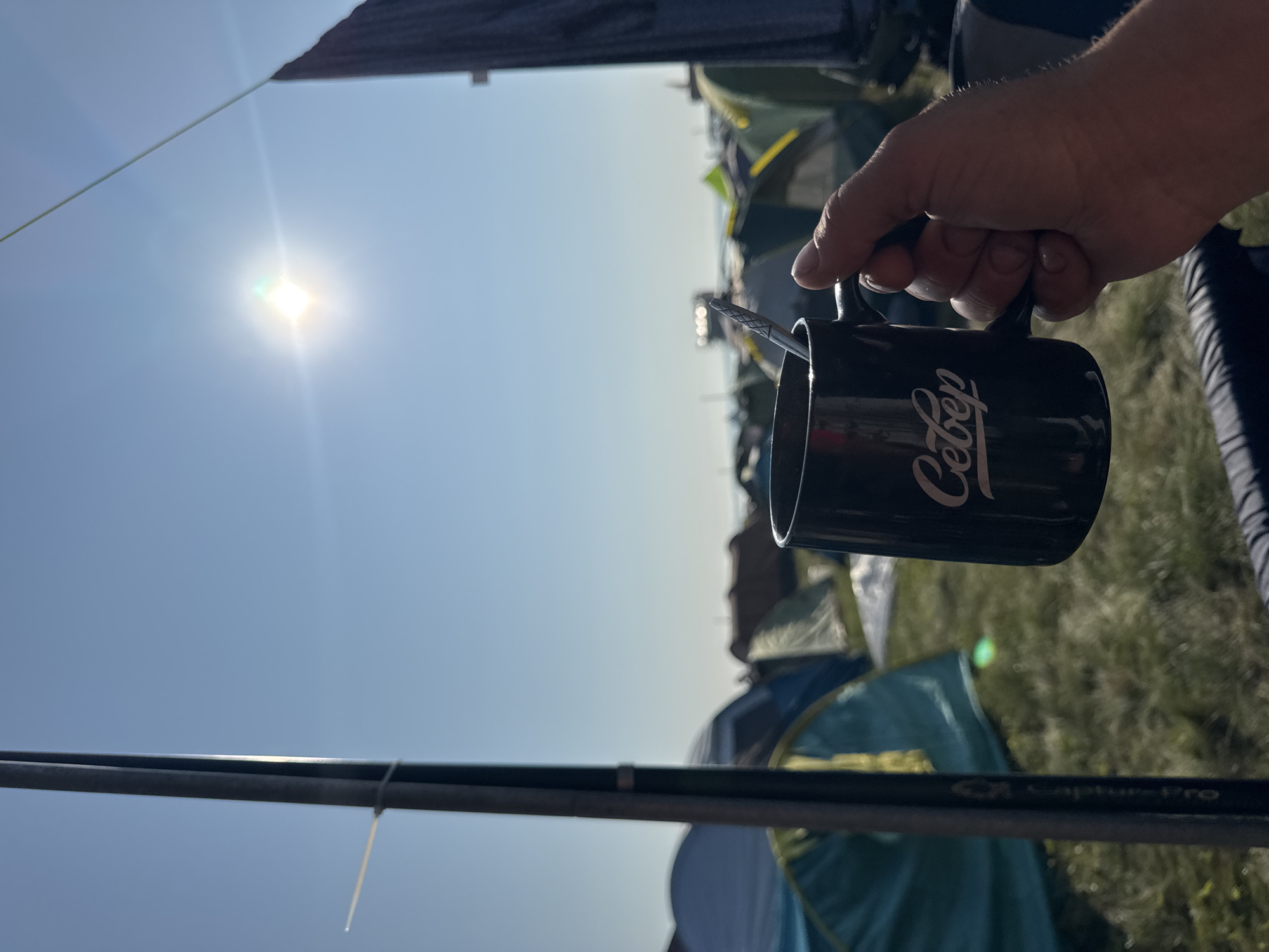 A first-person perspective shot captures a person holding a black mug with a silver spoon inside. The mug has the white Cyrillic word "Север" (Sever) printed on it. The background reveals a campsite under a clear, bright blue sky, with the sun high and prominent, casting a lens flare. Numerous tents of various colors, predominantly green and blue, are visible, suggesting an outdoor gathering or festival. A tall, dark pole is situated to the left of the frame, with a thin white cord attached to it. The ground is covered with grass.