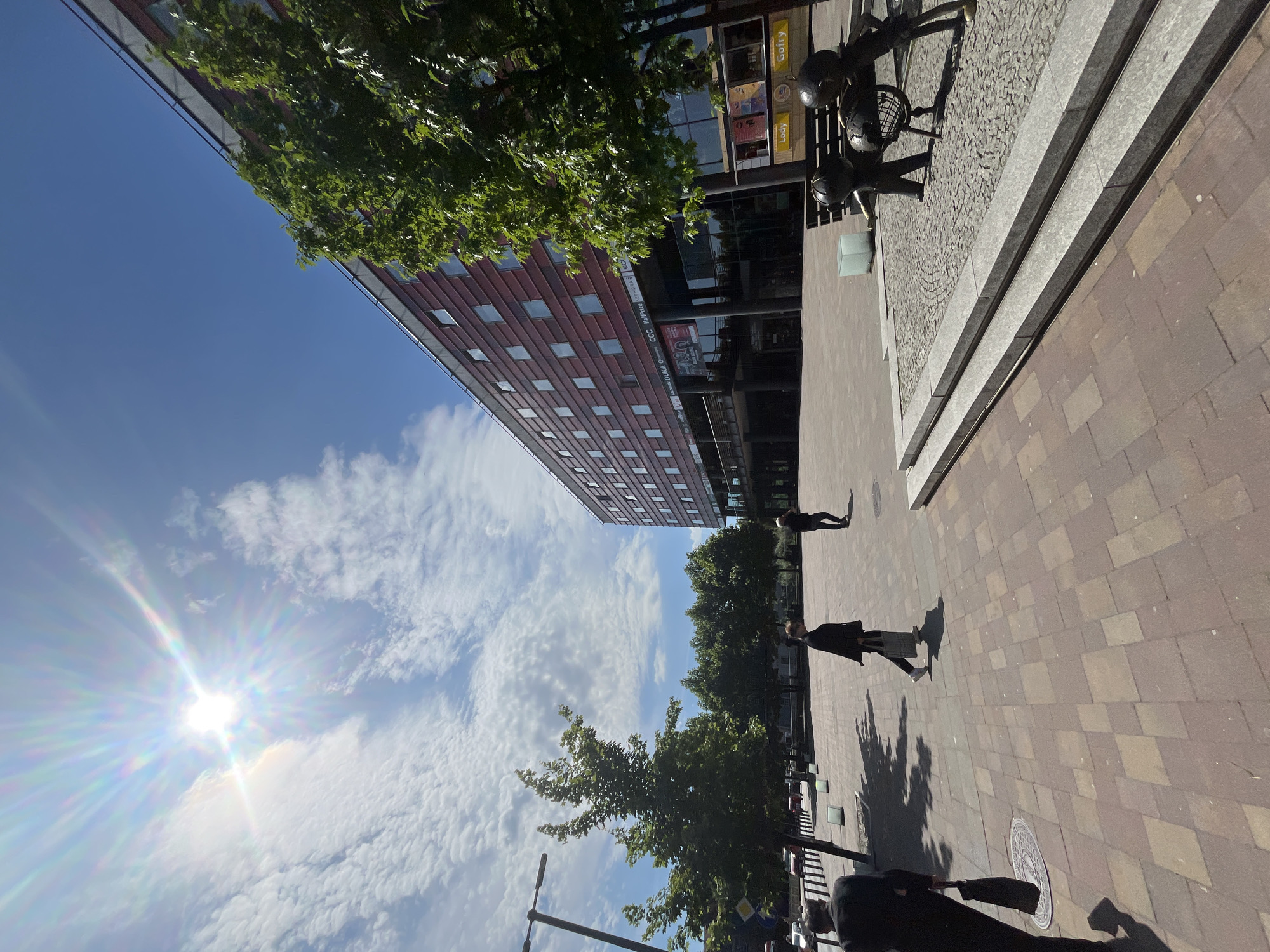 The image depicts an outdoor urban scene under a bright, sunny sky with scattered clouds. A modern, multi-story building with a red facade and numerous square windows dominates the right side of the frame. In the foreground, a paved plaza with brickwork patterns is visible. Several people are walking across the plaza. To the left, a lamppost stands. In the middle ground, there are trees and a low wall with a water feature. In the far right foreground, there is a bronze sculpture of two figures interacting with a globe. A bench is situated behind the sculpture. The sun is prominently visible in the upper left corner, casting lens flares.
