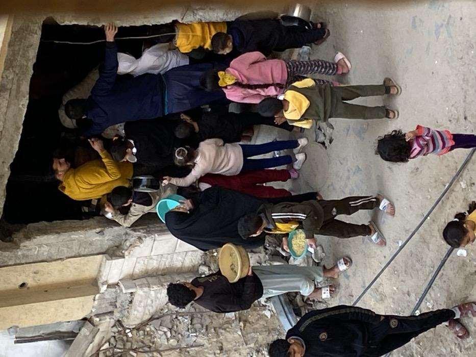 The image depicts an outdoor scene in Khan Yunis, Palestinian Territory, characterized by a gathering of people in front of a partially destroyed building. The structure on the left exhibits significant damage, with concrete rubble and exposed building materials. A large, dark opening, possibly a doorway or a section of a collapsed wall, serves as a focal point.
Approximately 20-25 individuals are visible, comprising numerous children and several adults. In the upper-central area, an adult male in a yellow hoodie stands inside the dark opening, observing the crowd. Another adult male in dark blue attire is also positioned within the opening. To the left, an adult male in a dark top and light pants holds a large yellow bowl containing a light-colored substance, likely food. An adult female, clad in a black abaya and light blue head covering, stands near the damaged building, holding a light blue plastic bucket.
Many children are clustered around the dark opening, some reaching upwards. A child in a green and yellow tracksuit in the mid-ground holds a yellow bowl filled with a light-colored food item. Other children are scattered throughout the foreground and mid-ground, some standing or looking around. The ground surface is composed of dirt and scattered debris, with visible electrical cables or ropes lying across it in the lower foreground. The overall scene suggests a distribution or collection of resources, likely food, amidst a damaged urban environment.