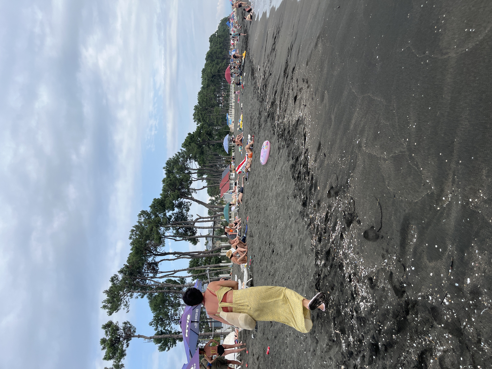 The image depicts a crowded black sand beach in Ureki, Georgia. In the foreground, a woman with short dark hair is walking away from the camera, wearing a yellow floral maxi dress and pink-and-white patterned slip-on shoes. She is carrying a large, beige, woven tote bag over her right shoulder. The beach is populated with numerous individuals lounging on towels and beach chairs under various umbrellas, some of which are purple and red. Several people are visible in swimsuits, some sunbathing, others sitting. The sand is dark and appears to have scattered shells and debris. Pine trees line the background of the beach, with some low-lying buildings visible behind them. The sky is overcast with patches of blue peeking through. The overall scene is one of a busy beach day with people enjoying the seaside.