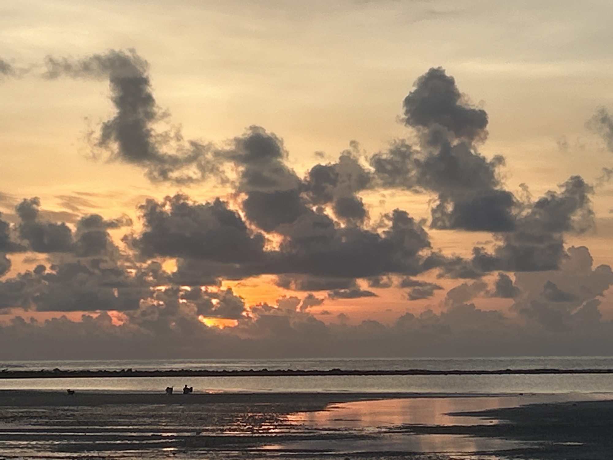 The image depicts a sunset at Baan Harn, Thailand. The sky is filled with grey clouds with an orange glow from the setting sun peeking through. Below the clouds is the ocean, a dark line separating the sky from the beach. The tide is low, revealing a wide expanse of sand. The sand is wet, with reflections of the sky visible in the water. Several people are visible in the distance on the beach, but they are too far away to discern specific details about their actions or identities.