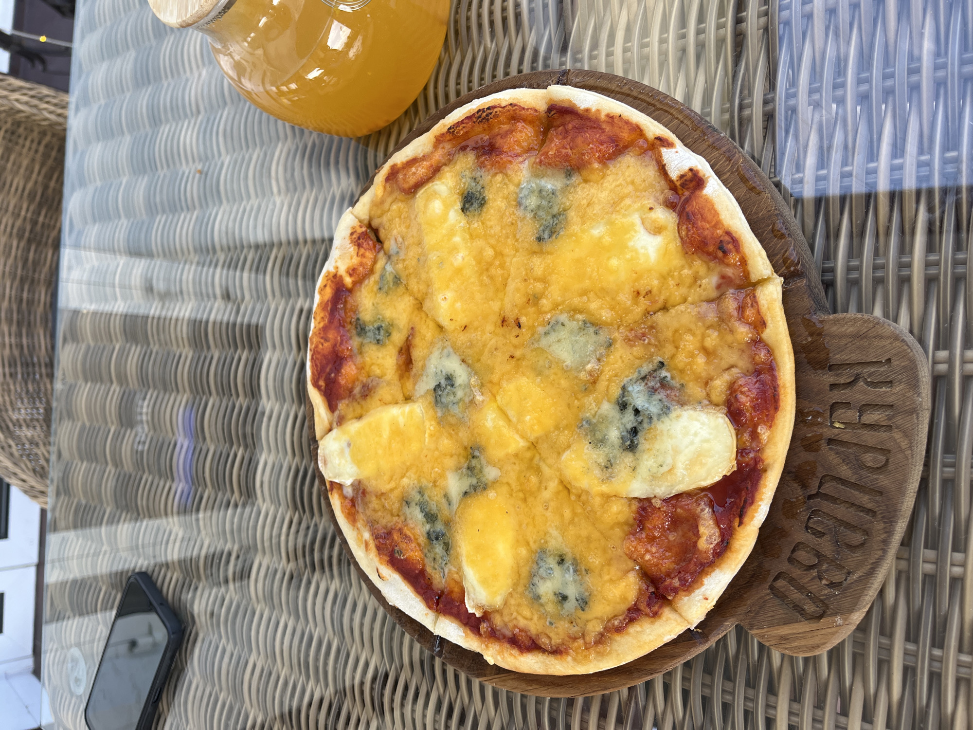 A close-up overhead shot shows a small, round pizza on a wooden cutting board with an inscription. The pizza is topped with melted cheese and red sauce. To the right of the pizza, a glass jar filled with an orange beverage sits on a woven table. A smartphone rests on the table to the left, partially obscured by the pizza. The scene appears to be outdoors, with the woven pattern of the table and wicker chairs suggesting a restaurant or cafe setting. The image is taken in Akhlebaikha (Kovriginskii S/S), Russia.