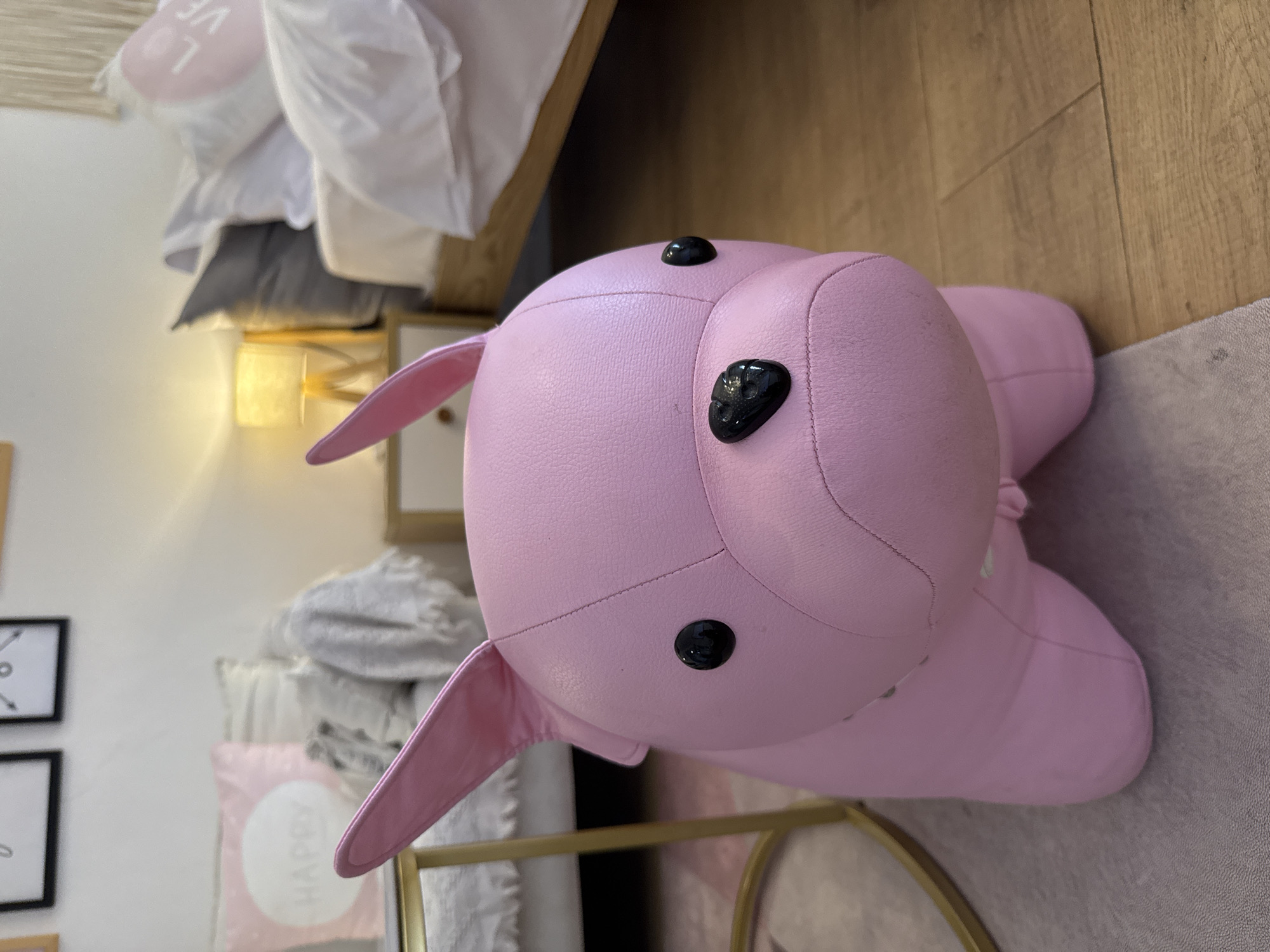 A close-up shot shows a pink, leather-like plush pig toy with black button eyes, lying sideways on a light grey rug indoors. A wooden floor is visible in the background. To the left, a bed with white bedding and a wooden frame is seen. A bedside table with a lamp on it, and other decor are also visible. The location is Jinjiang District, China.