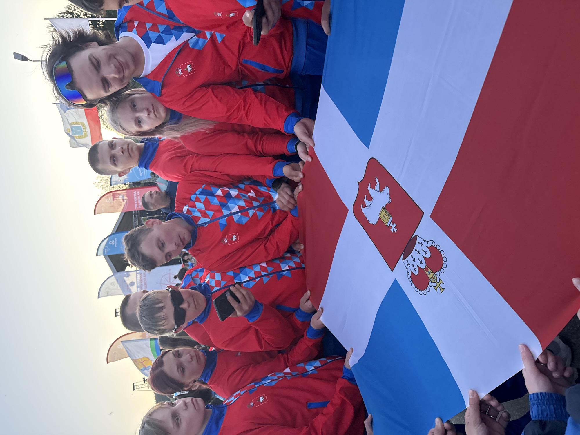 A group of people stands holding a flag that is divided diagonally into blue, white, and red sections. The flag has a crest on the white section. The people are wearing matching red jackets with blue and white detailing. One person is holding a smartphone, and another is wearing sunglasses. The background shows flags and what appears to be a stage or structure with signage. The setting seems to be outdoors, possibly during an event. Given the flag's colors and crest, the image is probably taken in Russia, maybe in Khvalynsk.