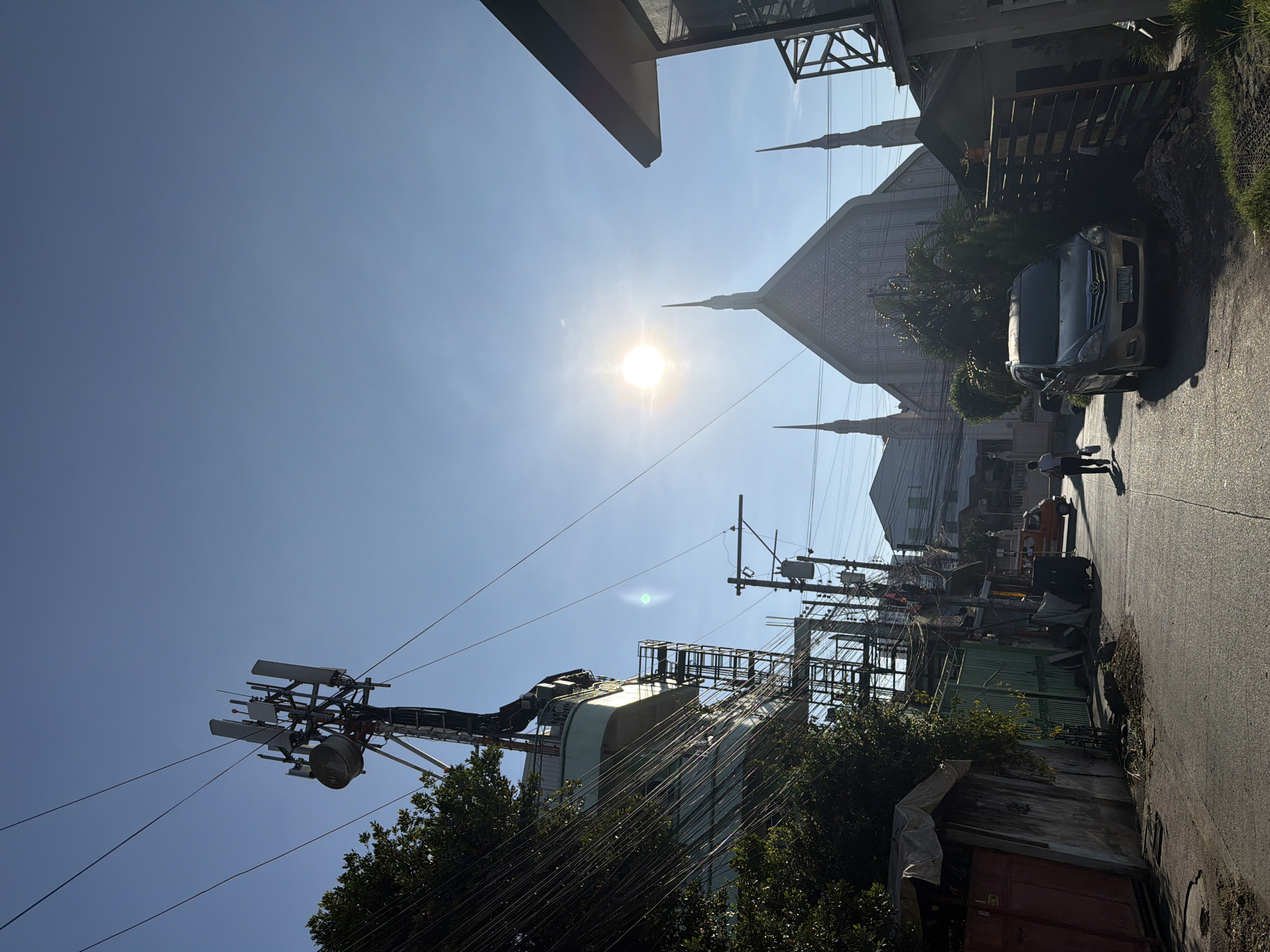 The image depicts a street scene in Davao City, Philippines, under a clear blue sky with the sun prominently visible. On the left, a tall cell tower stands amidst a tangle of electrical wires and lush green trees. A multi-story building with balconies and external staircases is also visible. Further down the street, on the right, a distinctive building with a triangular roof and spires, resembling a church or a similar religious structure, is situated. A silver SUV is parked on the side of the road in front of this building. A lone pedestrian, wearing a light-colored shirt and dark pants, walks away from the camera down the paved street, carrying a bag. The street is lined with various structures, including fences, buildings, and what appear to be utility poles with transformers. The overall impression is that of a sun-drenched urban environment with a mix of infrastructure and architecture.