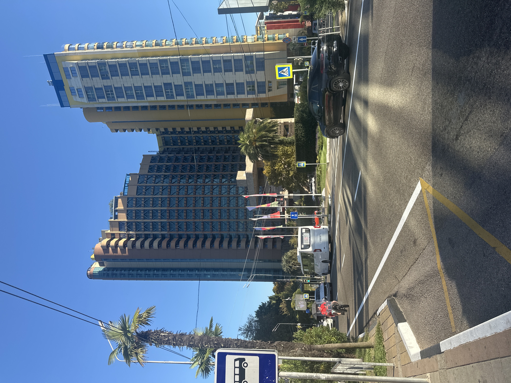This is a daylight, eye-level, exterior shot of a city street in Sochi, Russia, with a blue sky. In the foreground, a black SUV is turning right onto a street with white and yellow road markings. To the left, a palm tree stands next to a bus stop sign and a sidewalk. Further down the street, a white bus is driving away from the camera, and a person on a scooter is heading towards the camera. In the background, two modern, tall buildings dominate the skyline, with a palm tree situated between them. Several flags are also visible on poles in front of the buildings. The environment appears to be a bright, sunny day.