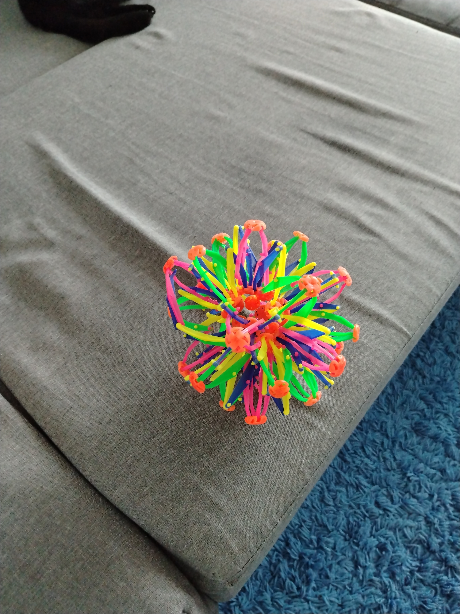 The scene is observed within a domestic interior, reportedly in the city of Смалявічы, Belarus. The central subject is a multi-colored, expandable kinetic toy, commonly identified as a Hoberman sphere, positioned on the edge of a grey upholstered furniture surface. The toy is composed of interconnected structural elements in neon green, bright pink, dark blue, and yellow, joined by distinctive orange spherical connectors. The grey upholstery, likely belonging to a sofa or cushion, displays a visible woven texture and minor creasing. In the upper left section of the image, a dark, amorphous object, possibly a piece of footwear, is partially visible and out of focus on the same grey surface. The lower right portion of the frame shows a shaggy, bright blue carpet or rug, providing a textural and chromatic contrast to the furniture. No human subjects or animals are present, and no overt actions or interactions are depicted. The lighting indicates an indoor setting.