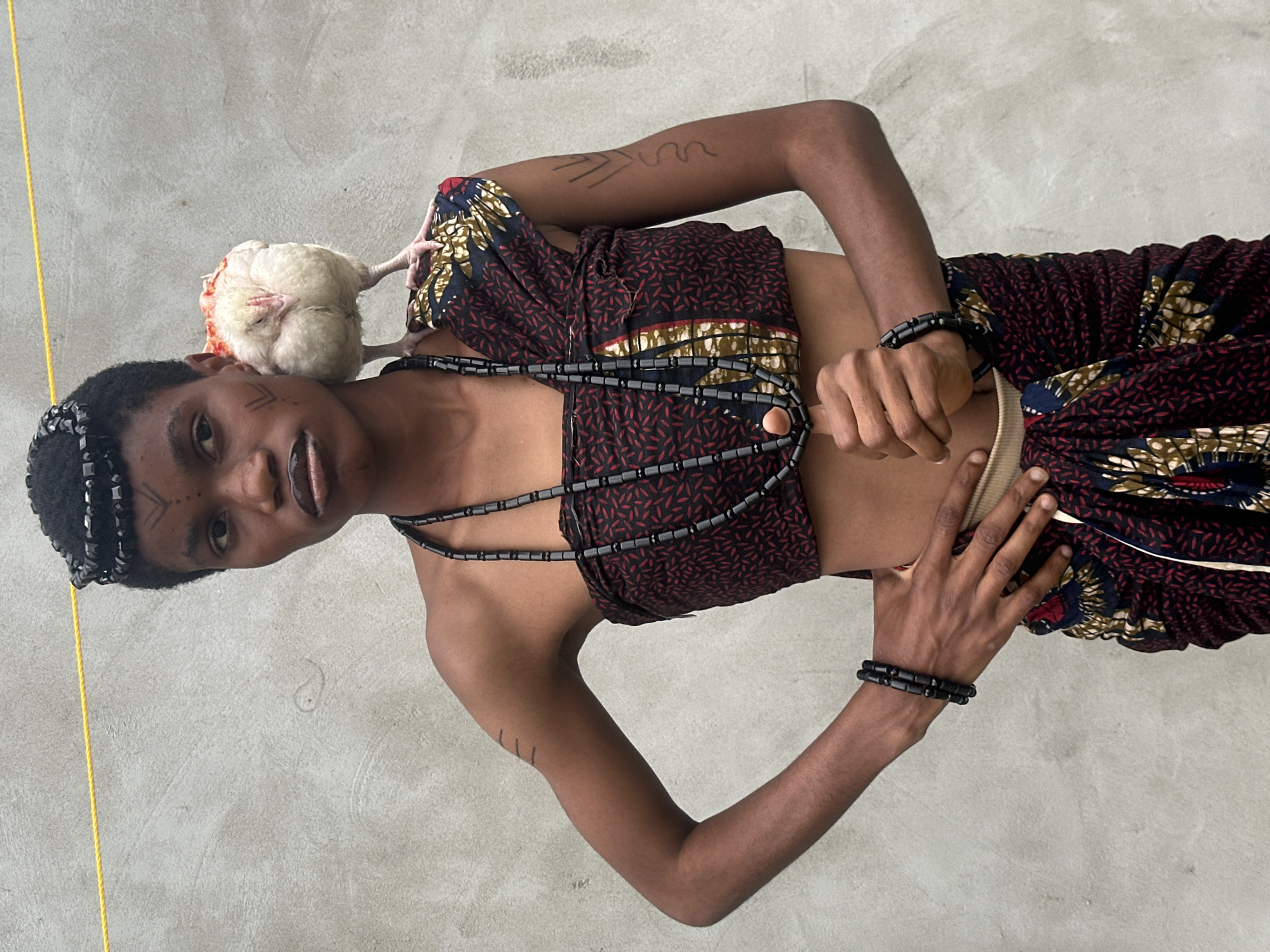 A young woman stands in front of a light gray wall, a yellow string visible at the top of the frame. She has dark skin and short, black hair, adorned with a black beaded headpiece. Her face is decorated with black tribal markings, including lines on her forehead and cheeks. She wears a patterned wrap top with red and black designs, and a matching wrap skirt. Around her neck, she has a long, black beaded necklace, and on her left wrist, a black beaded bracelet. A white chicken with red markings on its head is perched on her right shoulder. Her right hand is placed on her hip, while her left hand is slightly raised, fingers curled. The wall behind her appears textured and slightly weathered. The overall impression is one of cultural adornment and a connection with an animal.