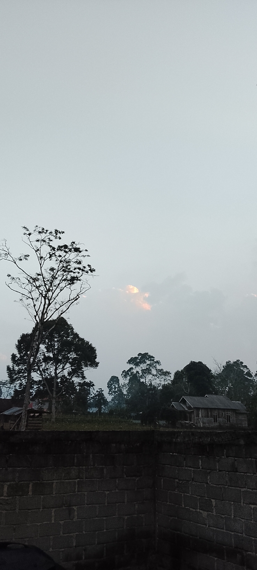A photograph captures a rural landscape at dusk in Padang Dalom, Indonesia. The foreground is dominated by a low, dark brick wall, with the corner of a building or structure visible on the right. Beyond the wall, a gently sloping field extends towards a line of trees and a few houses. The sky is overcast, with a soft, hazy light filtering through the clouds, hinting at the setting sun with a pale orange glow. Several trees, some bare-branched and others with foliage, are silhouetted against the sky. A simple, weathered wooden house with a sloped roof is visible amongst the trees on the right side of the frame, and a smaller, more dilapidated structure with a corrugated roof is present on the left. The overall atmosphere is quiet and serene, characteristic of a rural Indonesian village at the end of the day.