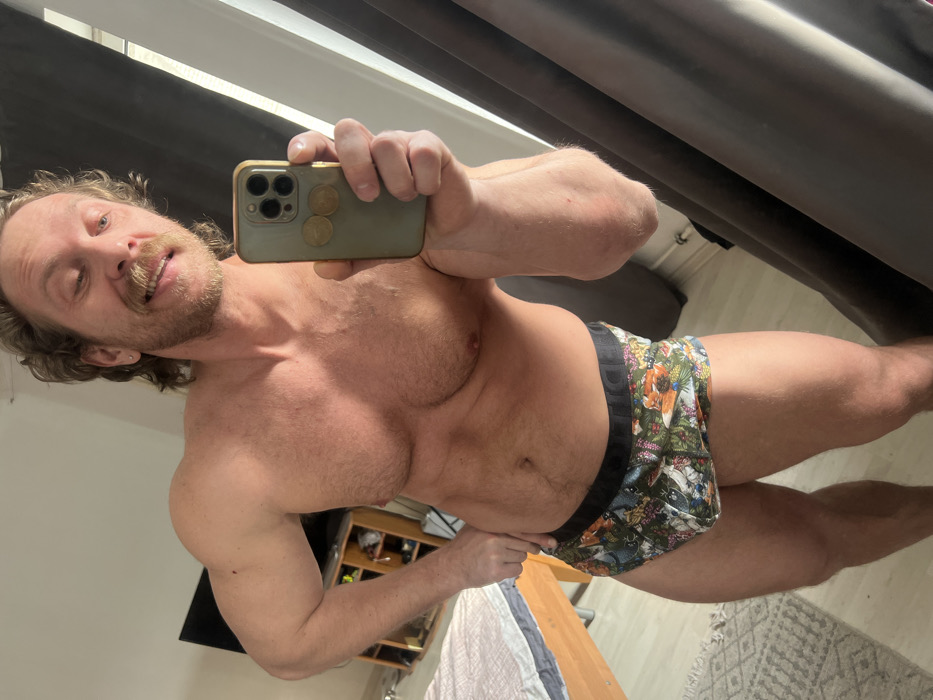 A male individual, appearing to be in his 30s or 40s, stands shirtless and wears colorful, patterned boxer briefs, captured in a mirror selfie. The subject possesses a muscular physique, light skin, light brown curly hair, a mustache, and a beard. He holds a light green smartphone with a gold-colored camera module and two circular, golden objects attached to its rear in his right hand, extended forward to capture the reflection. His left hand is positioned on his left hip. A slight smile is present on his face, with teeth partially visible. The background, visible in the mirror, shows an indoor setting, likely a bedroom or apartment. Elements include a wooden bed frame with a white headboard and a patterned comforter, a dark entertainment unit with a flat-screen television, a light-toned floor partially covered by a patterned rug, and dark gray curtains obscuring a window. The visible details do not provide sufficient information to forensically establish the specific location within the city of Saint Petersburg, Russia.