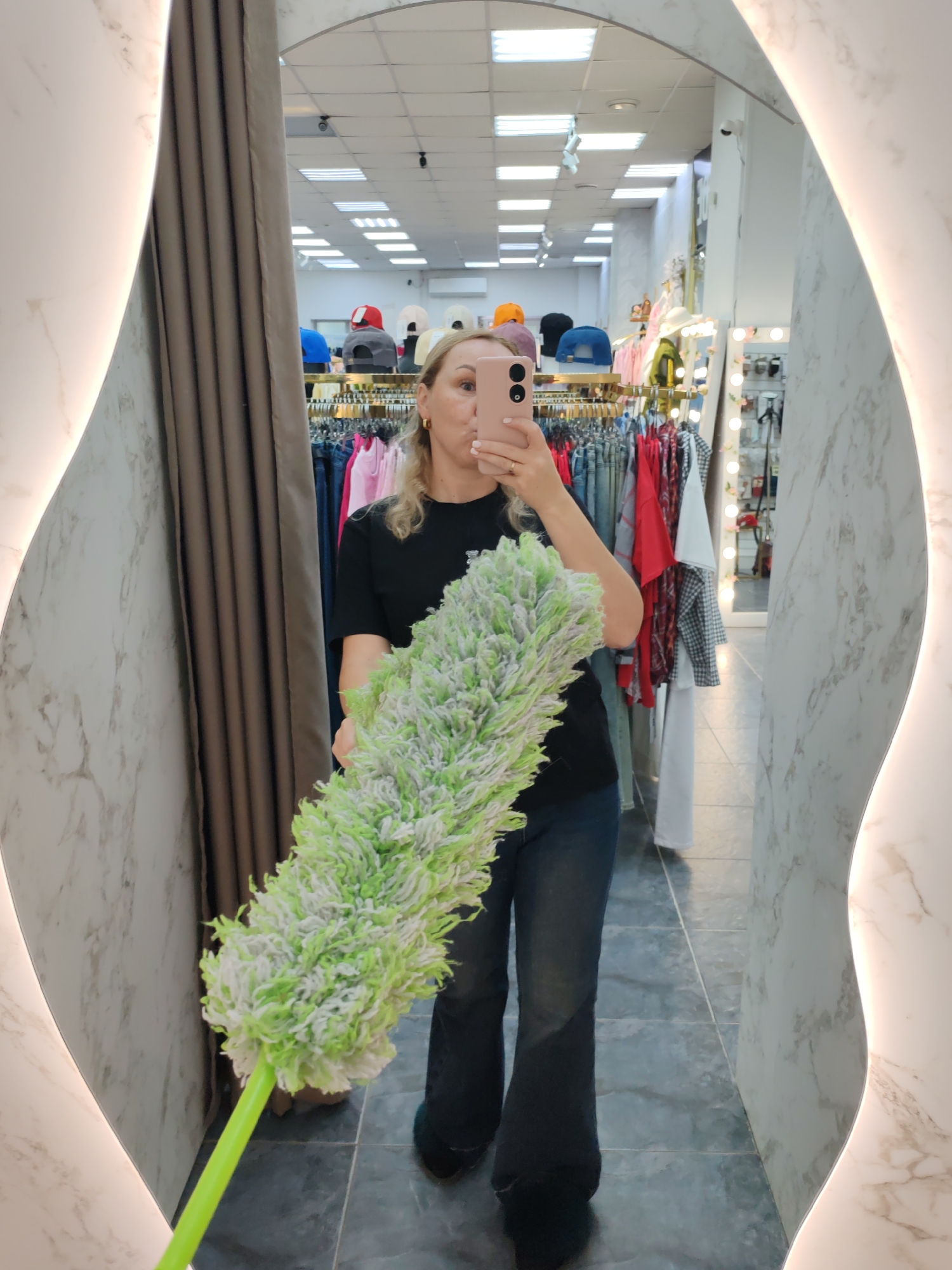 A woman is depicted taking a mirror selfie in what appears to be a retail clothing store or boutique. She is reflected centrally in a large, curved mirror with illuminated edges and a white and gray marble-patterned frame. The woman, who has blonde hair, is wearing a black t-shirt and dark blue jeans. She holds a light pink/beige smartphone to her face, obscuring most of her features, and her right hand shows a ring on her finger. In her left hand, she holds a prominent long, green-handled duster with a fluffy green and white head.
The background, visible through the mirror's reflection, shows a brightly lit store interior. Numerous clothing racks display various garments including t-shirts, jeans, and shirts in different colors. Above these, shelves hold a collection of hats in hues like red, blue, black, orange, and white. The ceiling features multiple fluorescent light fixtures and an air conditioning unit, while a security camera is mounted high on a wall. To the right of the woman's reflection, another mirror, possibly a vanity mirror with surrounding lights, is visible. A tall, brown curtain hangs to the left of the reflection. The floor is tiled. The scene suggests an everyday moment, possibly an employee taking a break or a customer playfully posing with an item for sale in Chaikovsky, Russia. No visible text is present in the image.