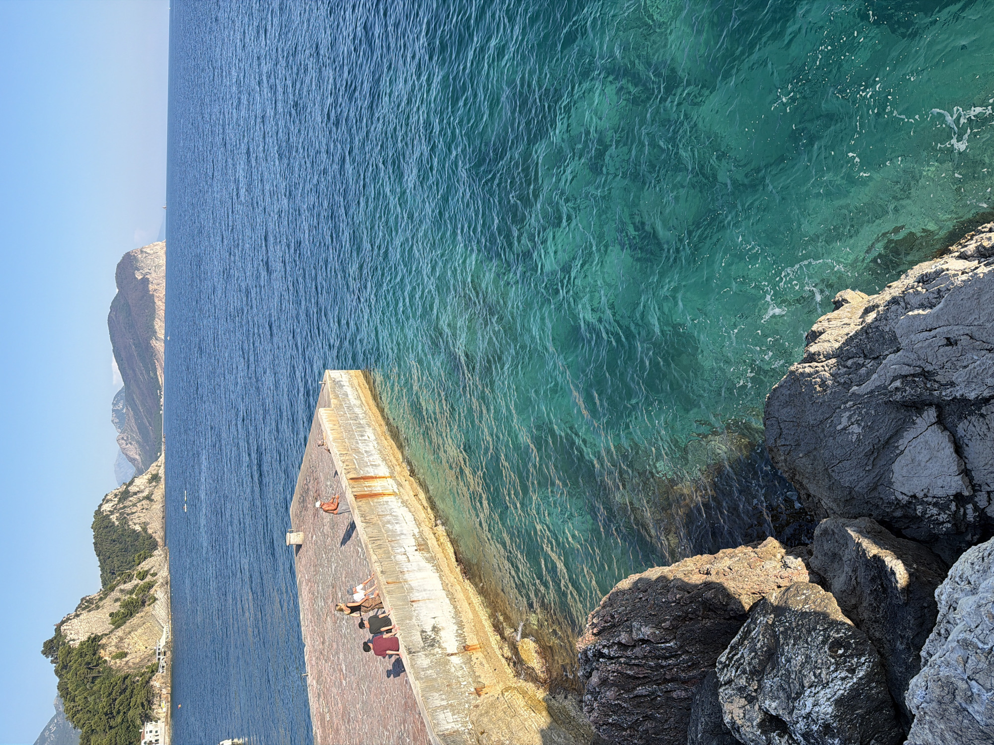 The image depicts a scenic view of the Adriatic Sea from a stone jetty in Petrovac centar, Montenegro. The clear, turquoise water is visible in the foreground, with sunlight glinting off its surface. The jetty, made of concrete and stone, extends into the sea. On the jetty, a small group of people are present. One person, a man in a white shirt and orange shorts, appears to be fishing. Several other individuals are seated or standing on the edge of the jetty, seemingly relaxing or observing the view. In the background, lush green hills and mountains rise from the coastline. The sky is a clear, bright blue, indicating a sunny day. The overall scene is tranquil and picturesque, characteristic of a coastal Mediterranean setting.