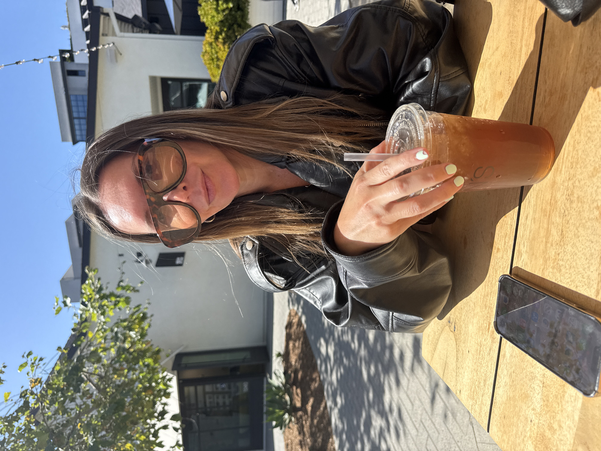 A woman is seated outdoors, looking directly at the camera with a relaxed expression, in what appears to be a cafe or patio setting in San Diego, United States of America. She has long, straight brown hair and wears large, tortoise-shell sunglasses and a black leather jacket. Her nails are painted a light pastel green.
She is holding a clear plastic cup filled with an iced, amber-colored beverage, likely iced tea or coffee, with a straw. A faint initial, possibly "S" or "JS", is visible on the side of the cup. Her left hand holds the cup, with her fingers wrapped around it.
The woman is seated at a light-colored wooden table. To her right, a smartphone with a visibly cracked screen is lying on the table. Strong sunlight illuminates the scene, casting distinct shadows on the table and ground, indicating a bright, clear day, likely late morning or early afternoon.
In the background, there are modern buildings with white and dark grey facades under a clear blue sky. Some greenery, including trees and shrubs, is visible. String lights are strung overhead in the distance, suggesting a decorative outdoor ambiance. The overall impression is one of leisure and relaxation in a sunny urban environment.