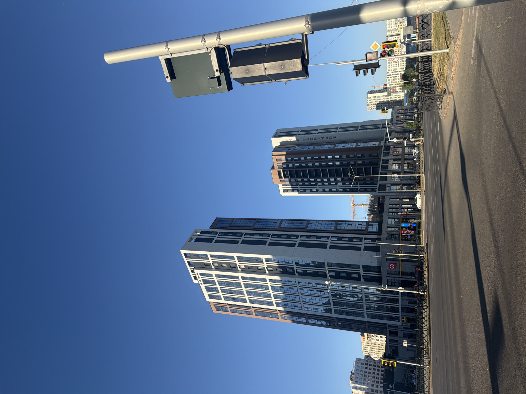 The image shows a street scene in Astana, Kazakhstan, featuring modern high-rise buildings under a clear blue sky. In the foreground, a paved road with lane markings is visible, casting long shadows from an unseen light source, indicating it's likely morning or late afternoon. A traffic light pole stands prominently on the right side of the image, equipped with multiple traffic signals and a sign.
Two distinct tall buildings dominate the midground. The building on the left is a modern residential or office tower with a facade composed of white, dark gray, and reddish-brown panels, interspersed with numerous windows. The text "CREST PLAZA" is visible near its peak. To its right, another similar but slightly shorter building has a facade of dark gray, white, and some brown elements, with the text "PROMENADE" vertically displayed along its side. In the background, more residential buildings of varying heights and architectural styles are present, along with a construction crane, suggesting ongoing development in the area.
Various vehicles are observed on the road and parked along the street, including cars and a white sedan. Several pedestrian crossing signals and traffic lights are positioned at an intersection. The ground level of the buildings features commercial storefronts, one of which displays a "60" sign, possibly indicating a speed limit or a store name. The overall environment appears to be a developed urban area with contemporary architecture.