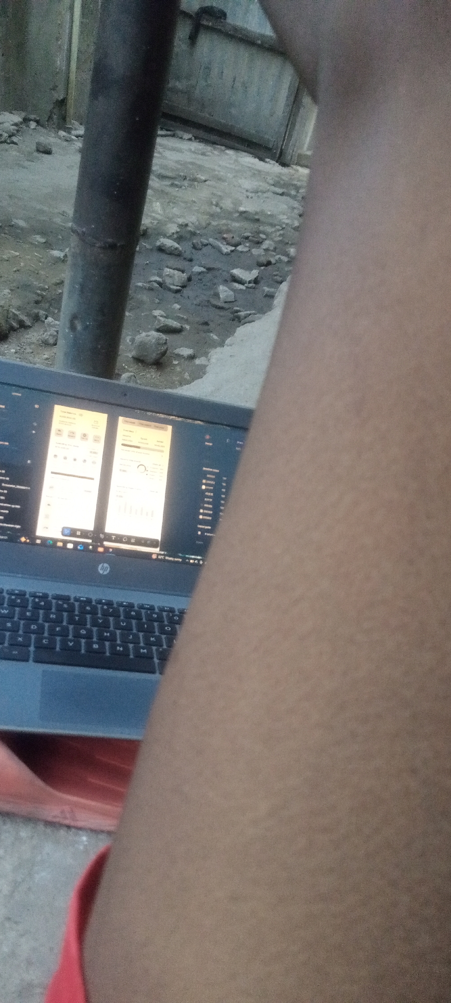 The image shows a person sitting outdoors in Yelwa, Nigeria, using an HP laptop. The laptop screen displays two windows with graphs and data. The person's bare leg is prominently featured in the foreground. A metal pipe is visible in the background, along with a concrete structure and rough ground with rocks. The person is wearing a red garment.