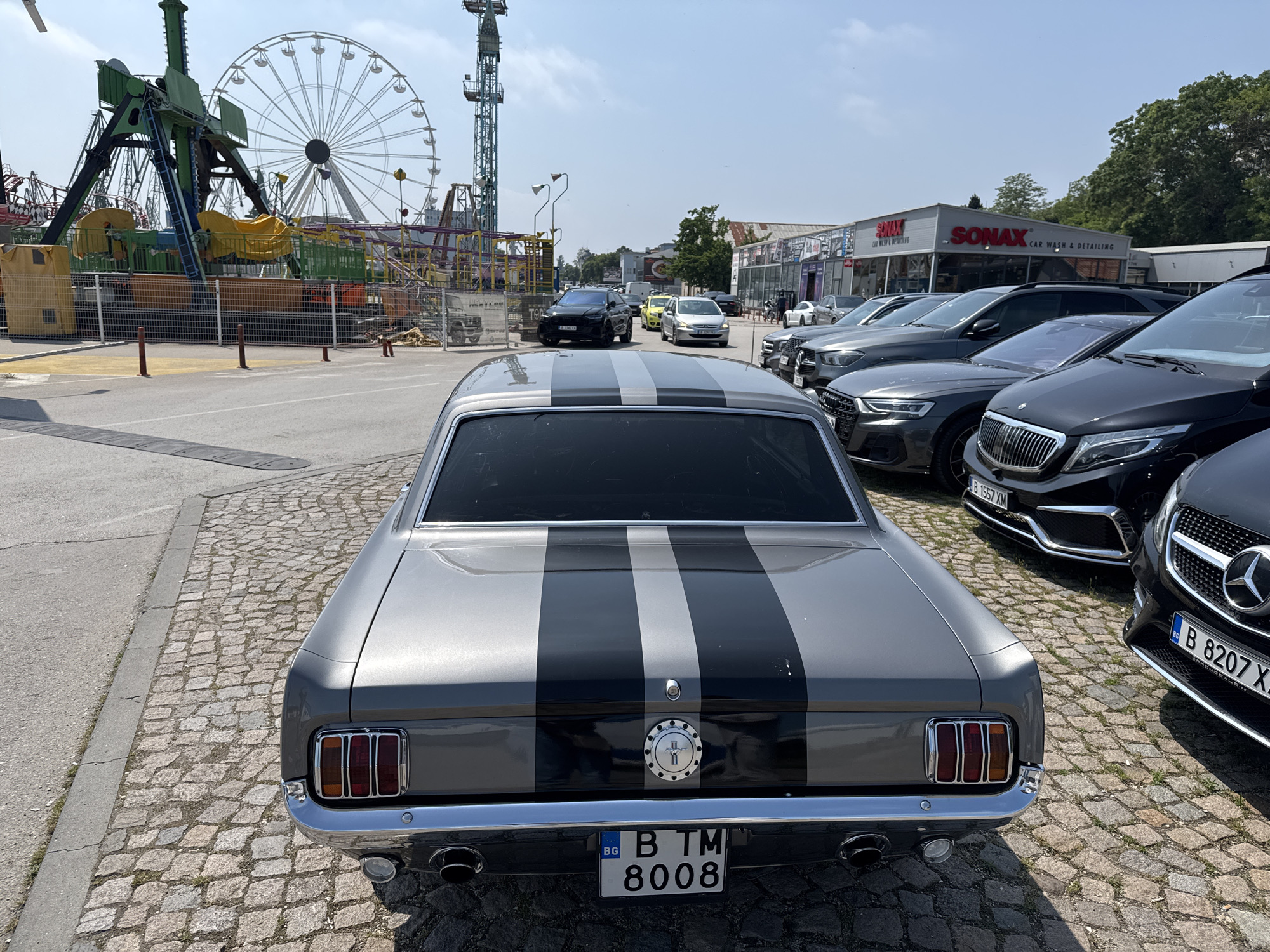 The image captures an outdoor scene dominated by a classic grey Ford Mustang with bold black racing stripes, viewed from the rear. The car is parked on a cobblestone surface, with its license plate reading "BG B TM 8008". Dual exhaust pipes are visible beneath the chrome bumper, and the iconic Mustang emblem adorns the trunk lid.
To the right of the Mustang, a line of modern, mostly dark-colored cars is parked on what appears to be an asphalt lot, including several Mercedes-Benz vehicles with license plates "B 1557 XM" and "B 8207 X.". Across the street from these parked cars, a commercial building with prominent red "SONAX CAR WASH & DETAILING" signage is visible.
In the background, to the left, an amusement park or fairground is evident, featuring a large white Ferris wheel and a tall green thrill ride, possibly a drop tower or swinging attraction. Fences and other amusement structures are also present. The ground transitions from cobblestones near the Mustang to asphalt roads and parking areas.
The scene takes place on a clear, sunny day, indicated by the bright lighting and lack of clouds in the blue sky. The overall setting appears to be an urban or semi-urban area, possibly a commercial district or entertainment zone, given the mix of car services and amusement rides. No people are explicitly visible in the frame, but the parked cars and commercial establishments suggest a functional and active environment.