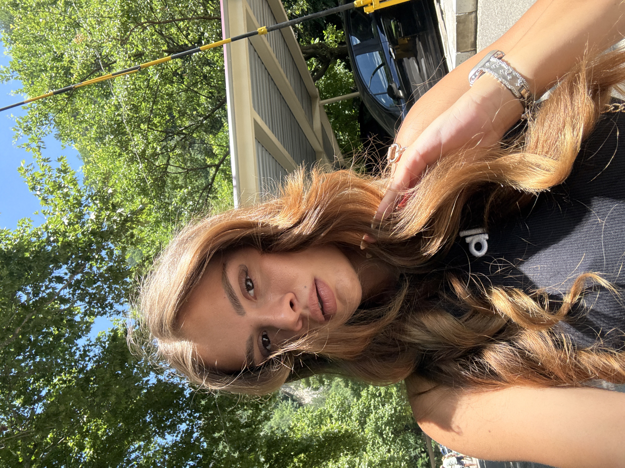 The image shows a young woman with long, wavy, light brown hair, taking a selfie outdoors. She is wearing a black tank top with a white logo and a silver watch on her left wrist. She is also wearing a ring on her left index finger. In the background, there are lush green trees, a bright blue sky, and a covered parking area with a black car parked beneath it. A yellow and black striped pole is visible on the right side of the image. The woman is outdoors during the daytime, with the sun casting shadows. The location is identified as Bachkovo, Bulgaria.
