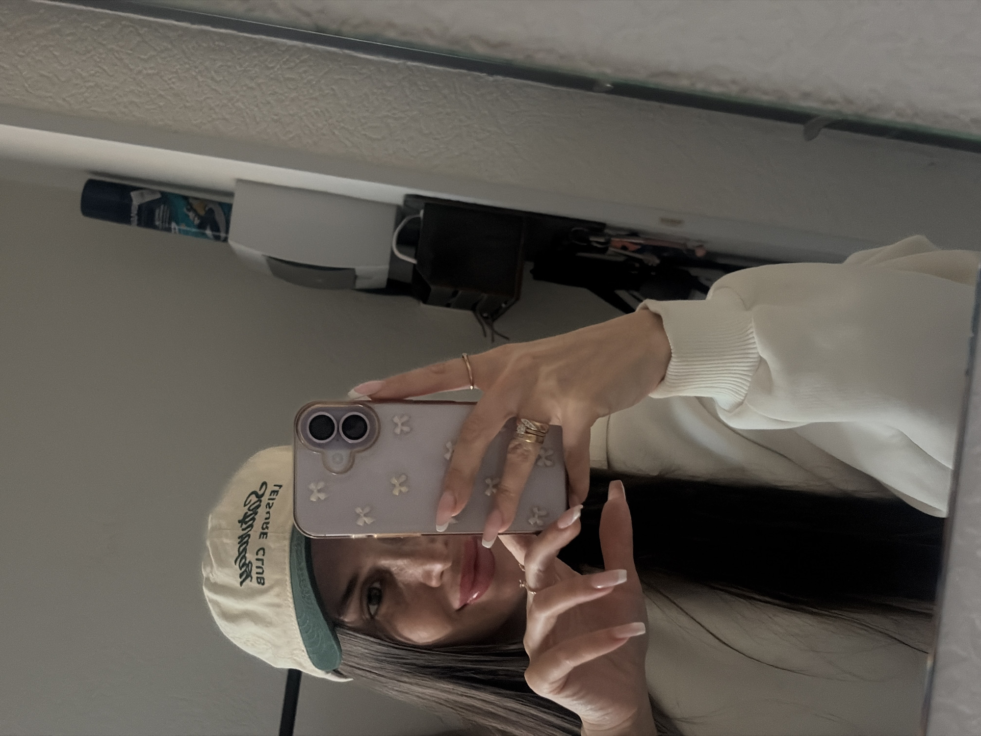 A person is taking a selfie in a mirror. The person is wearing a baseball cap with the words "Leisure Club" written backward. The person is also wearing a white sweater and has long, dark hair. The person is holding a cell phone with a light purple case and white flower designs. The background is blurry but appears to be a room with a light-colored wall and some sort of equipment mounted to it. There is no indication of the location being Saint Petersburg, Russia.