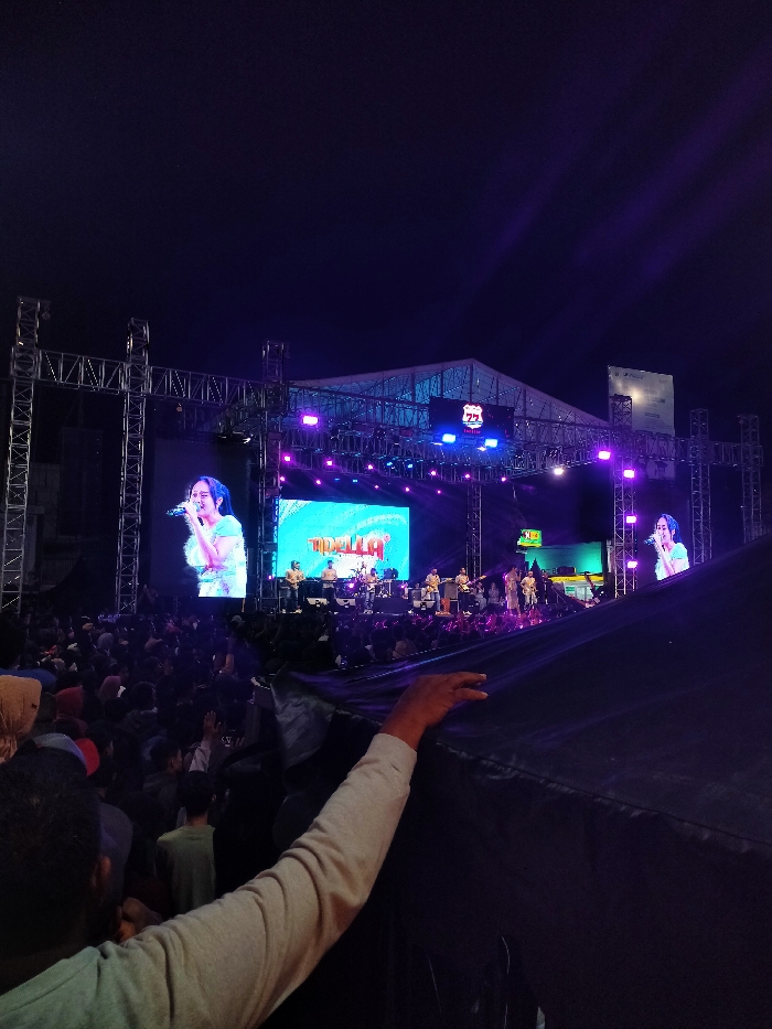 The image shows a concert taking place in Boyolali, Indonesia at the coordinates -7.535, 110.601. The stage is set up outdoors under a large white tent with a metal trussing system overhead. It is lit with colorful spotlights. A large screen is behind the stage featuring a woman singing. The stage is also populated with musicians, some playing instruments, some standing in front of microphones. The concert is taking place at night and a large crowd is gathered in front of the stage. Many in the audience are using their cell phones to record the performance. In the foreground, a person's hand reaches out from under a black cover toward the stage. The person's arm is outstretched towards the performance. The image appears to have been captured from behind the black cover. The concert appears to be a popular event.